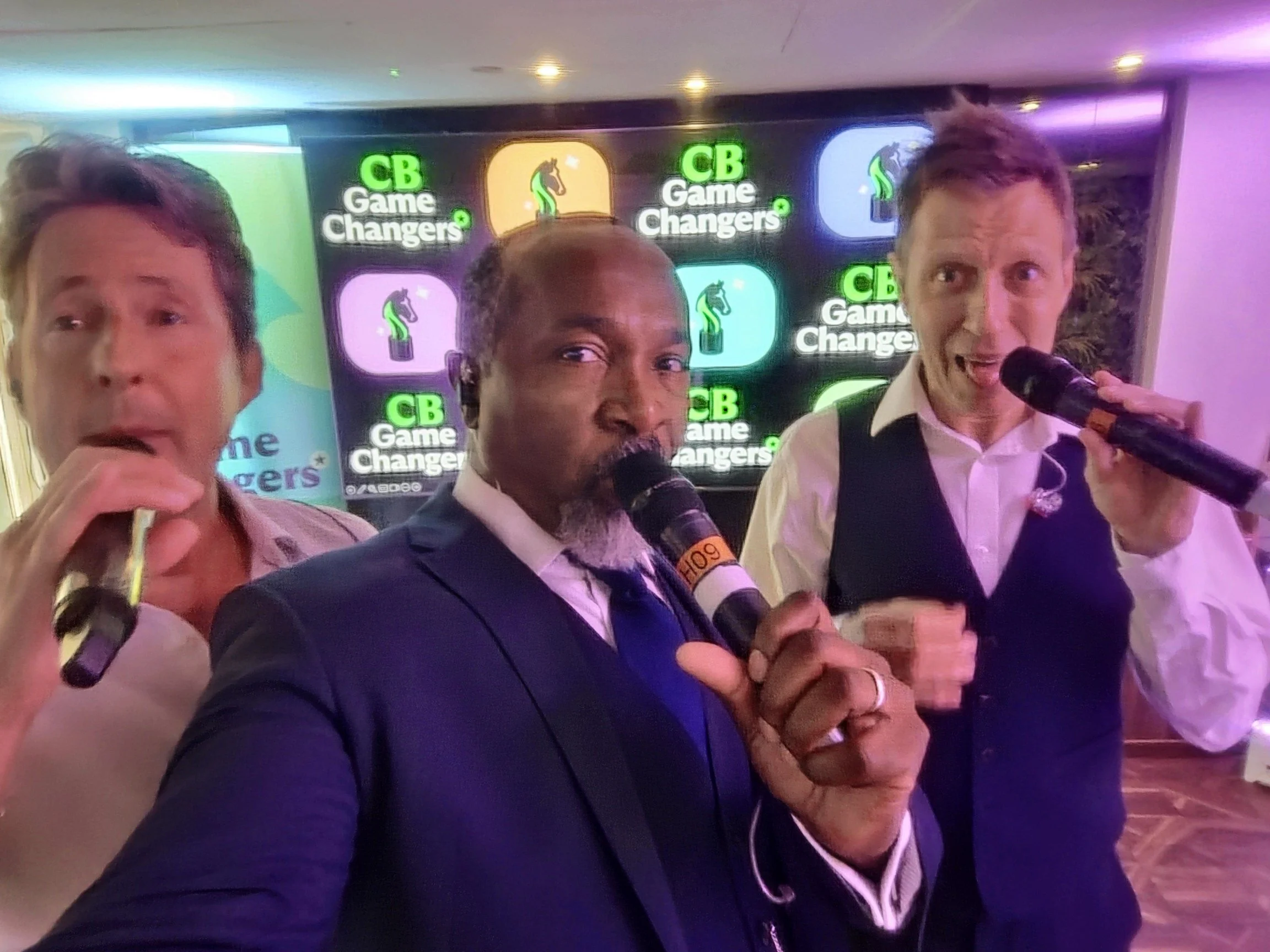 Three men singing into microphones at an event with a backdrop featuring the 'CB Game Changers' logo.