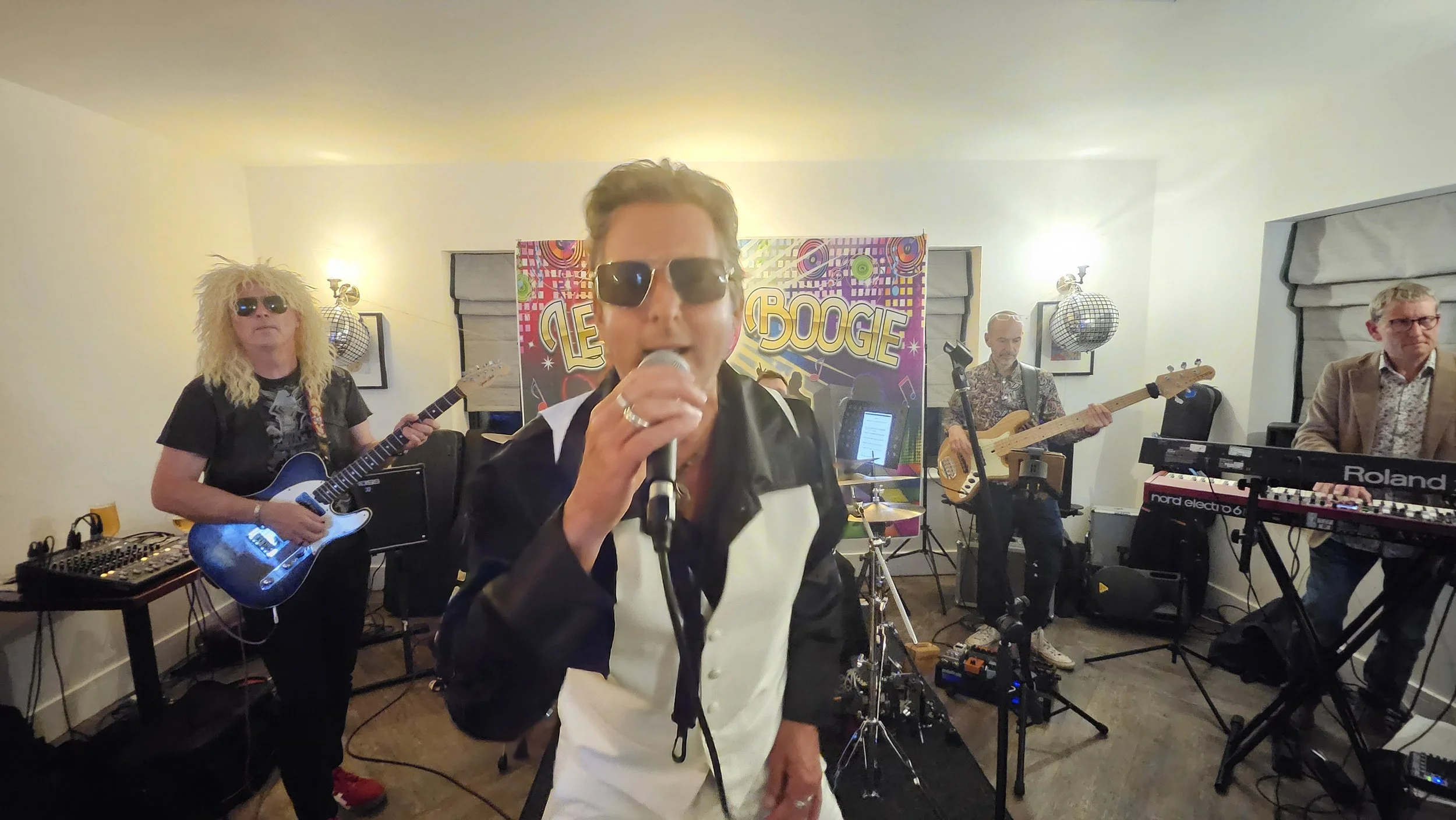 A band performing live at a party with a colorful sign that reads 'The Boogie'. The singer, in the foreground, wears sunglasses and holds a microphone. Other band members are playing guitar, bass, keyboard, and DJ equipment in the background.