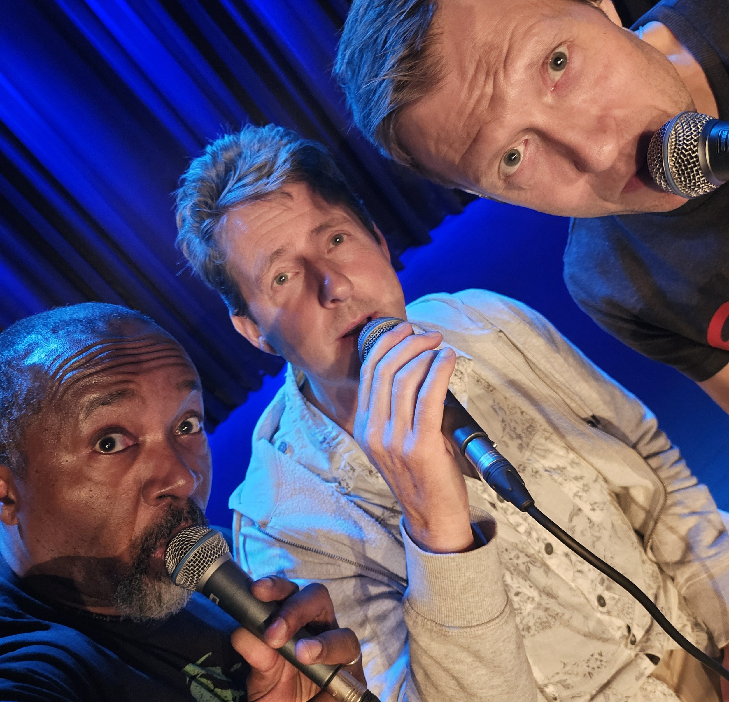 Three men close together, each holding a microphone; one with a gray beard and curly hair, one with short gray hair and light eyes, the third with dark hair, all appearing to be singing or speaking in a lively setting with dark blue background.