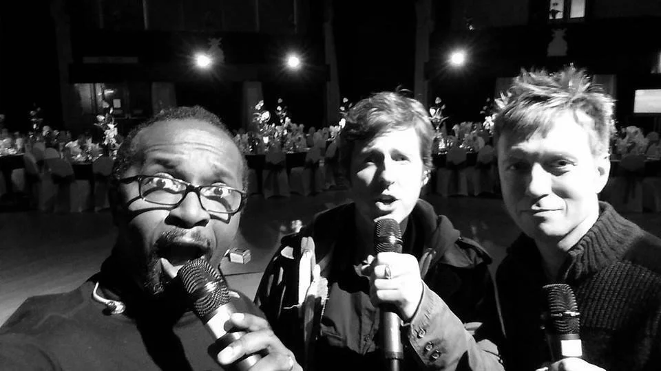 Three men holding microphones and taking a selfie in a dimly lit event hall with tables and seated people in the background.