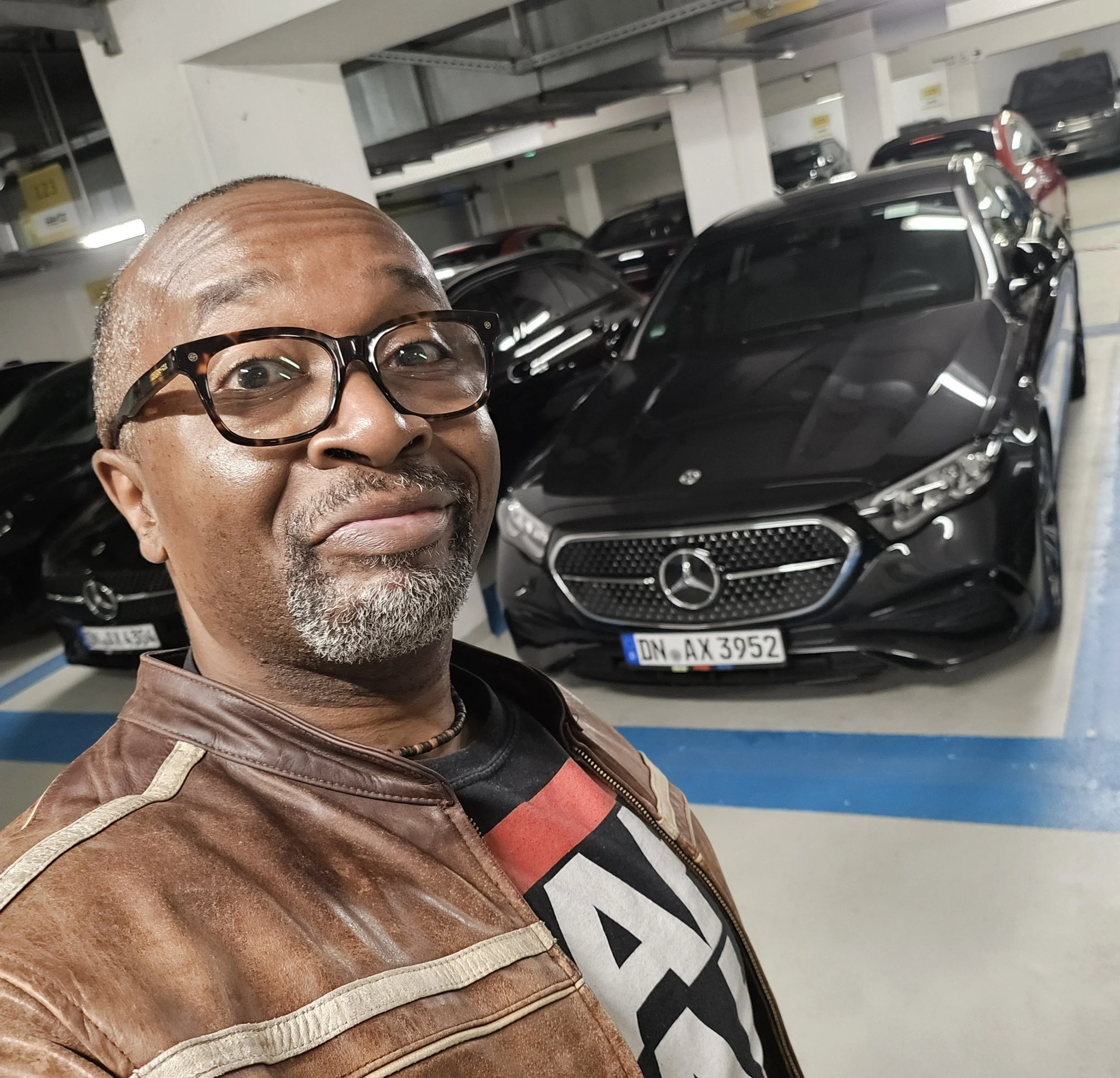 A close-up of a man with glasses, a bald head, gray beard, wearing a brown leather jacket, taking a selfie in an underground parking garage with black luxury cars in the background.