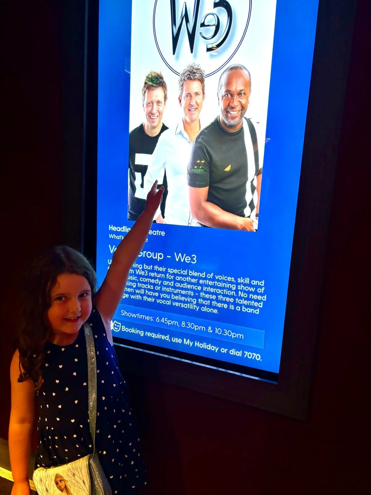 A young girl pointing at a digital display screen with a group photo of three men and information about a singing group called We3. The girl is wearing a navy dress with white polka dots and holding a bag featuring a doll or character from a movie.