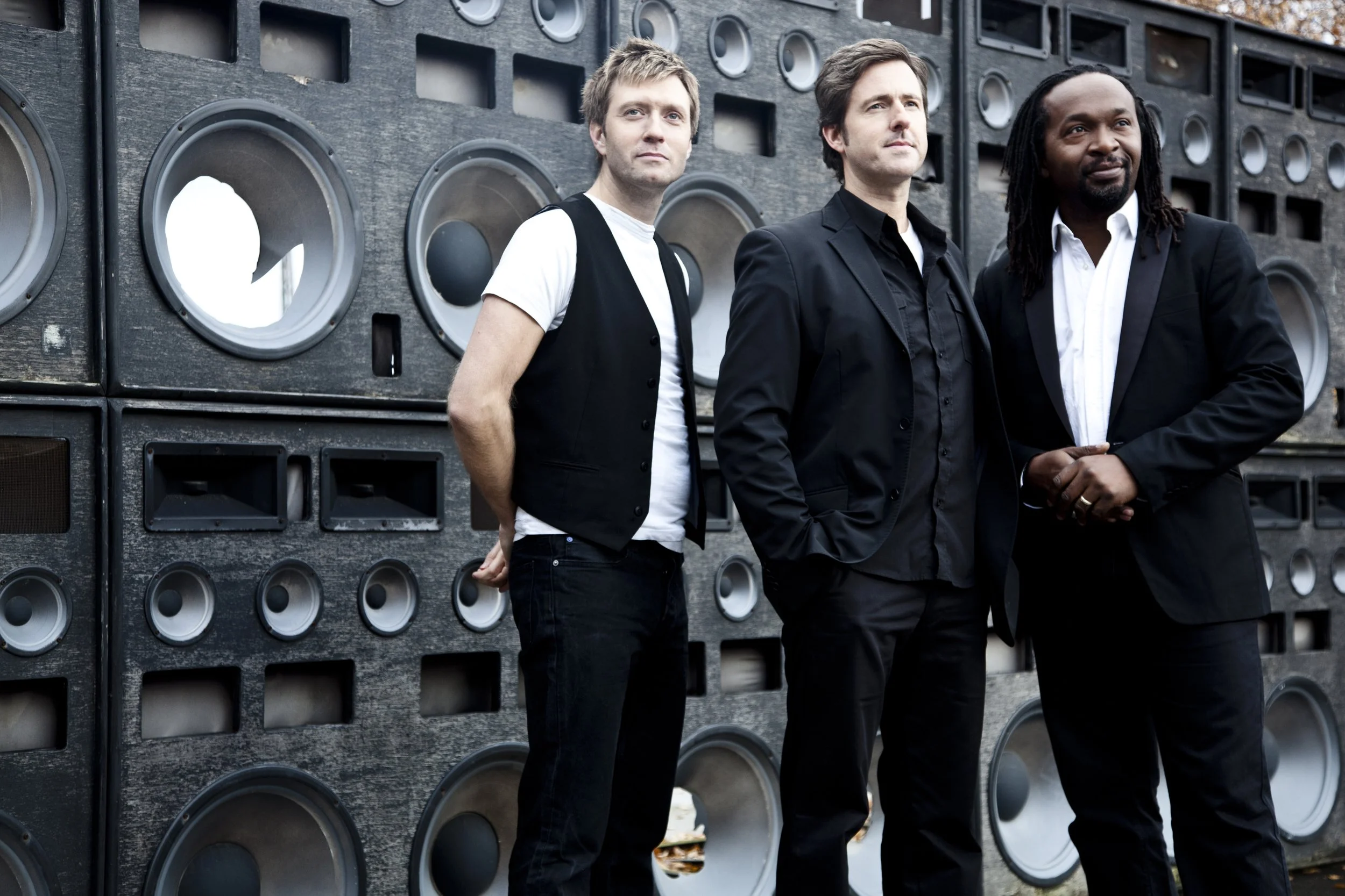 Three men standing in front of a large wall of vintage speakers. The men are dressed in black and white suits and looking confidently into the distance.