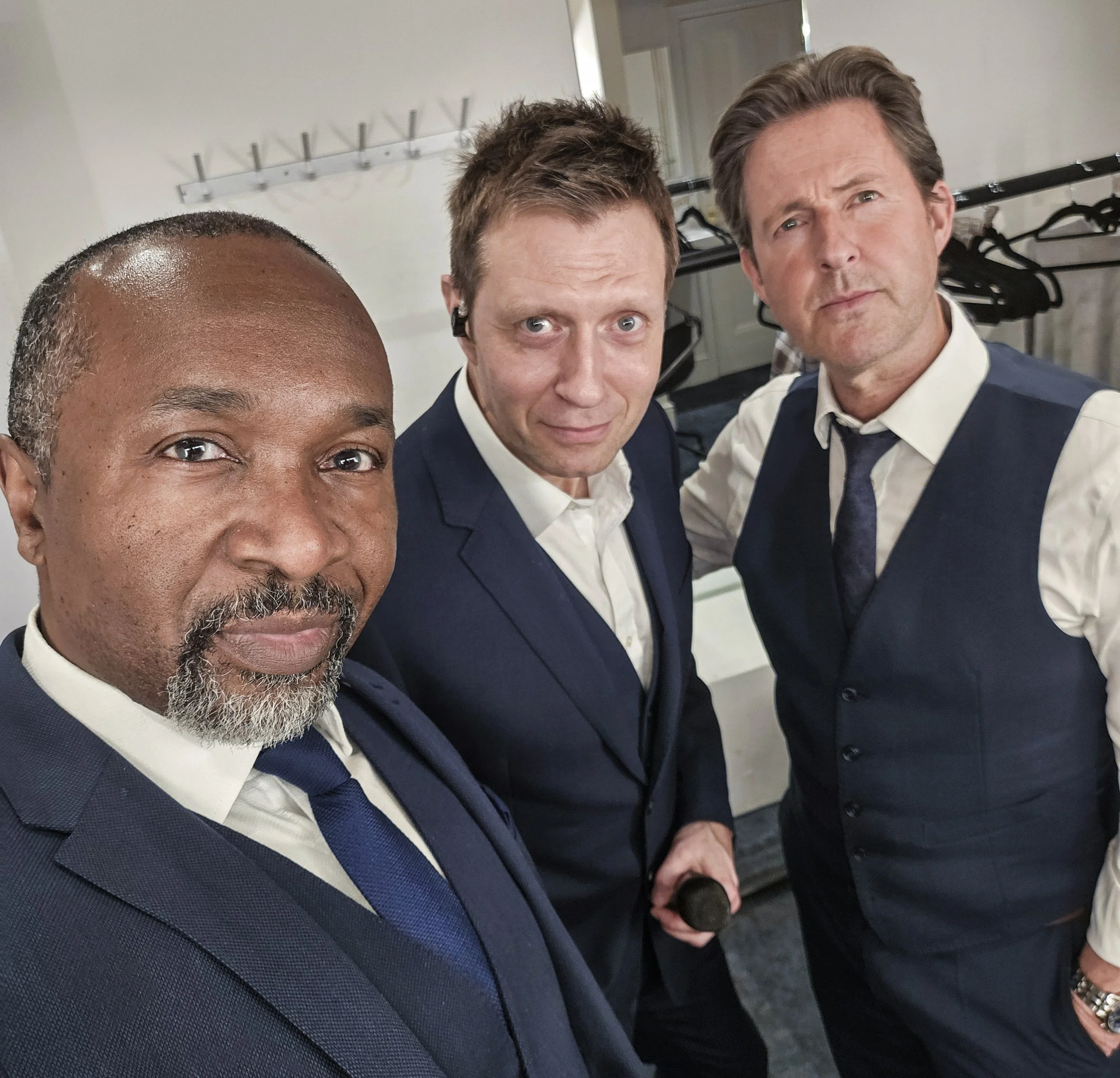 Close-up selfie of three men dressed in formal suits and white shirts, standing closely together and looking at the camera, in an indoor setting with coats hanging on a rack in the background.