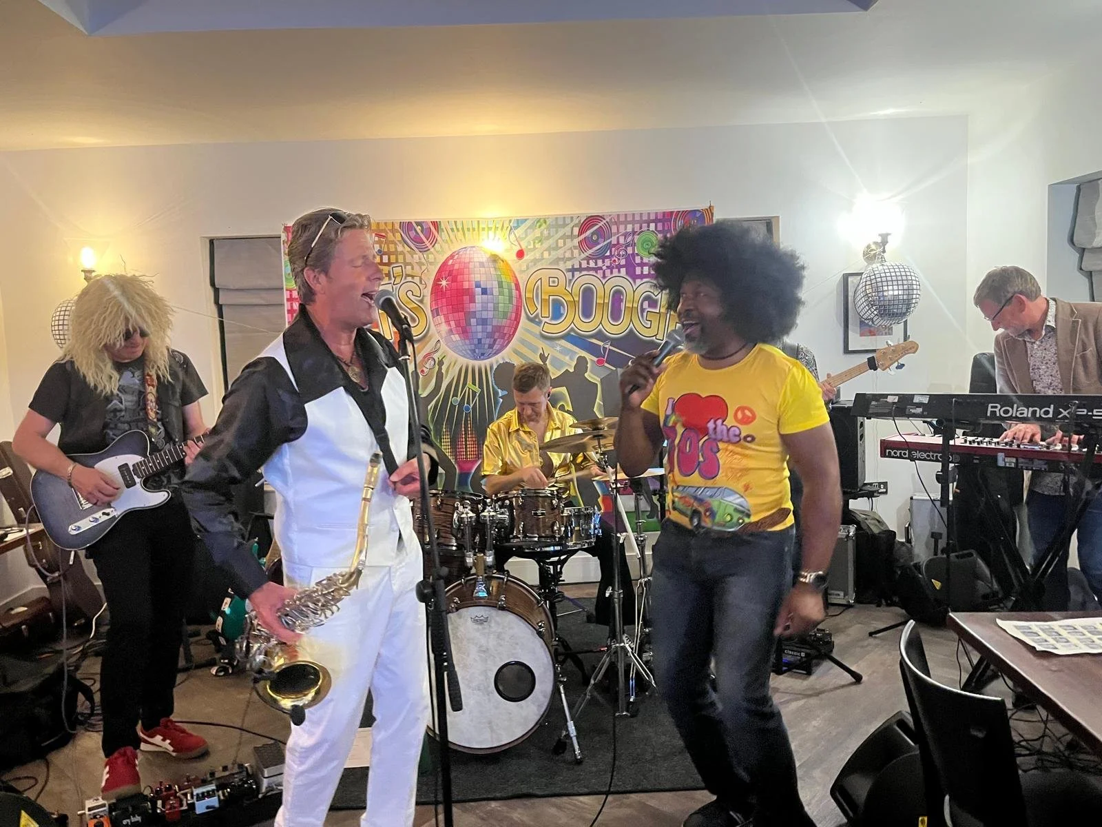 A band performing live on stage with six members, including a guitarist, two singers, a drummer, a keyboardist, and a bassist. The backdrop features a colorful disco ball and music notes.