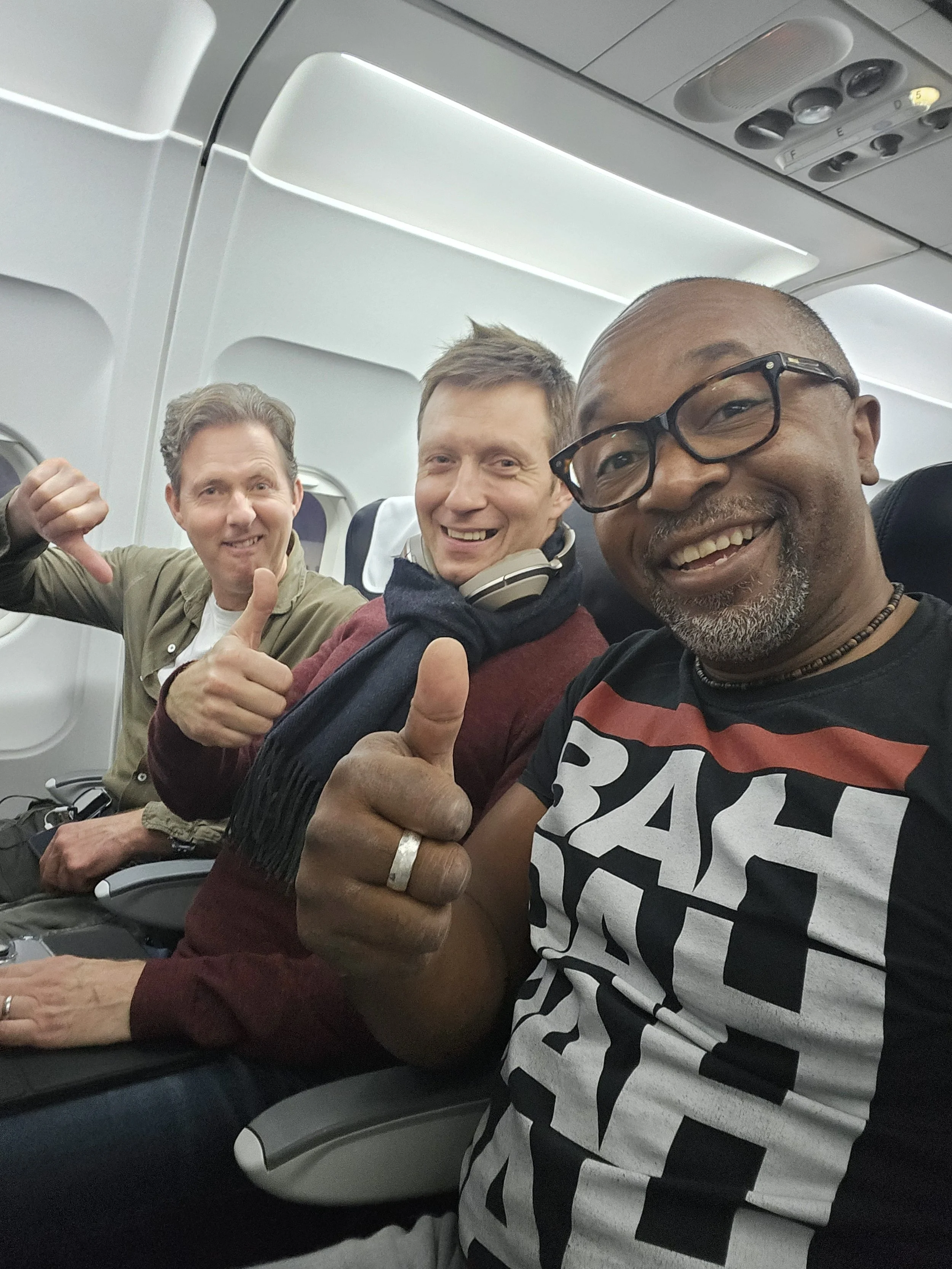 Three men sitting in airplane seats, smiling and giving thumbs up. One man on the right is wearing glasses and a black and white graphic t-shirt. The middle man has headphones around his neck. The man on the left is making a thumbs-down gesture.
