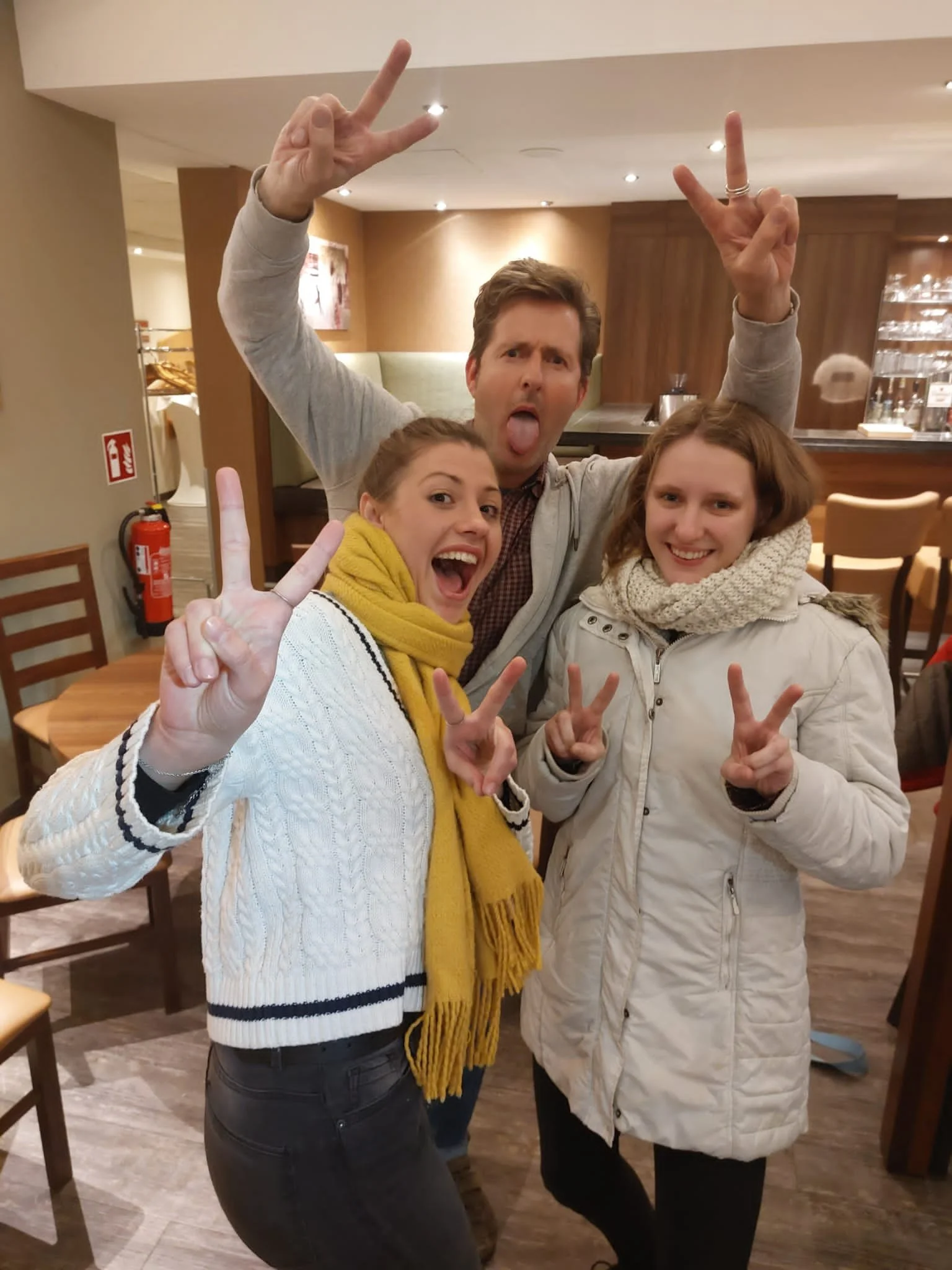 Three friends posing and making peace signs in a restaurant or cafe setting, with a man sticking out his tongue in the middle.