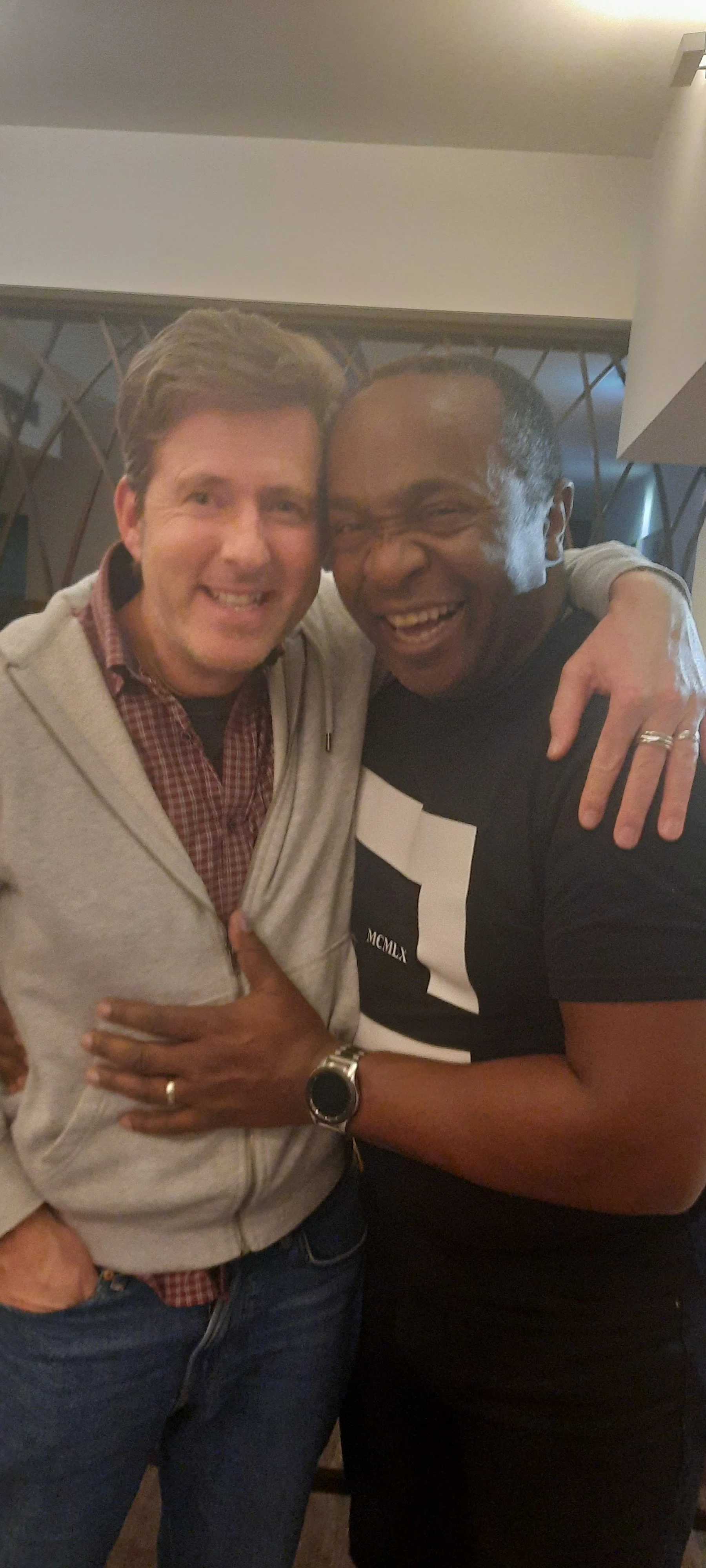 Two men smiling and hugging each other in a friendly embrace indoors.