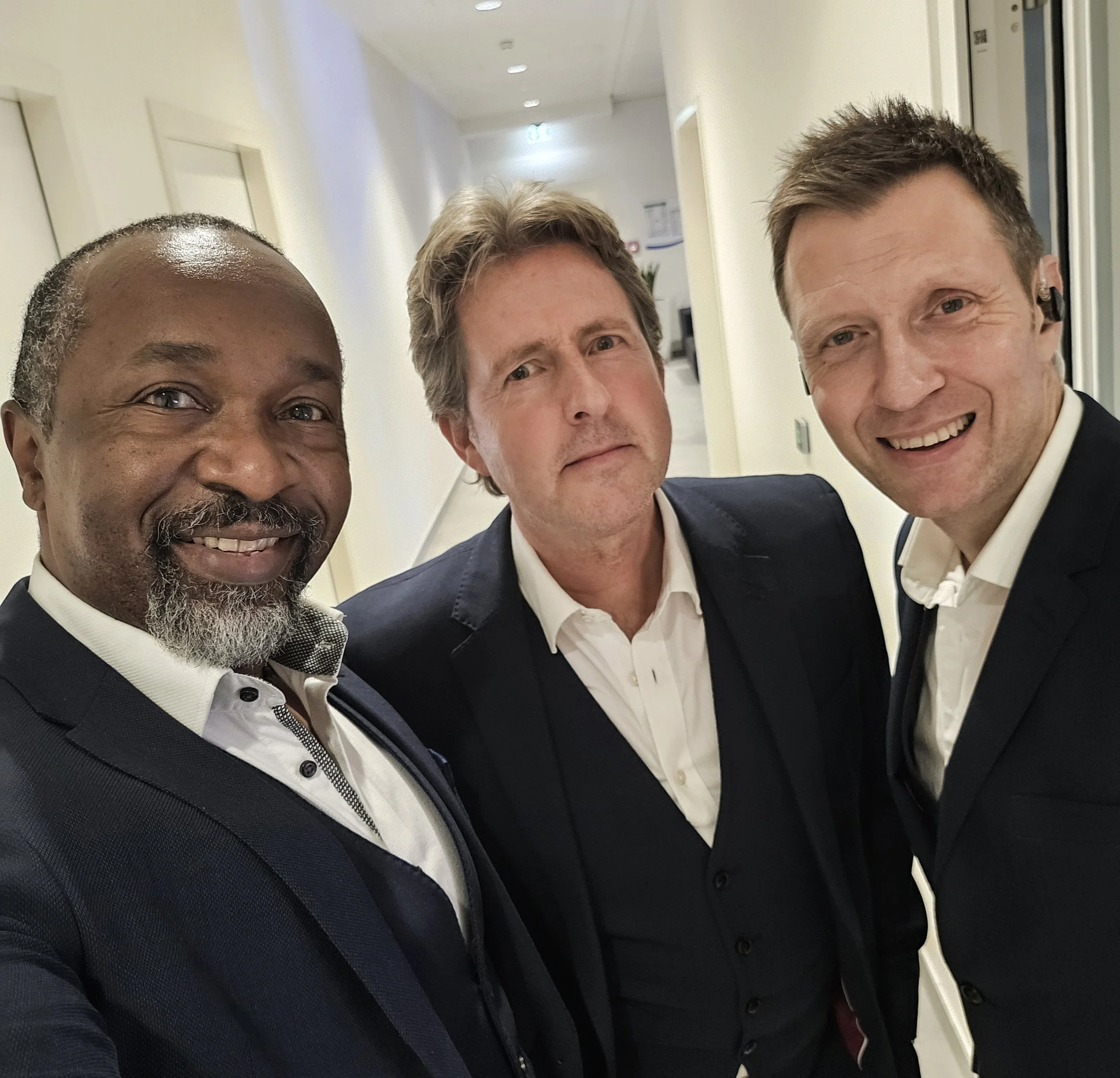 Close-up of three smiling men in suits standing close together in an indoor setting, possibly an office or conference area.