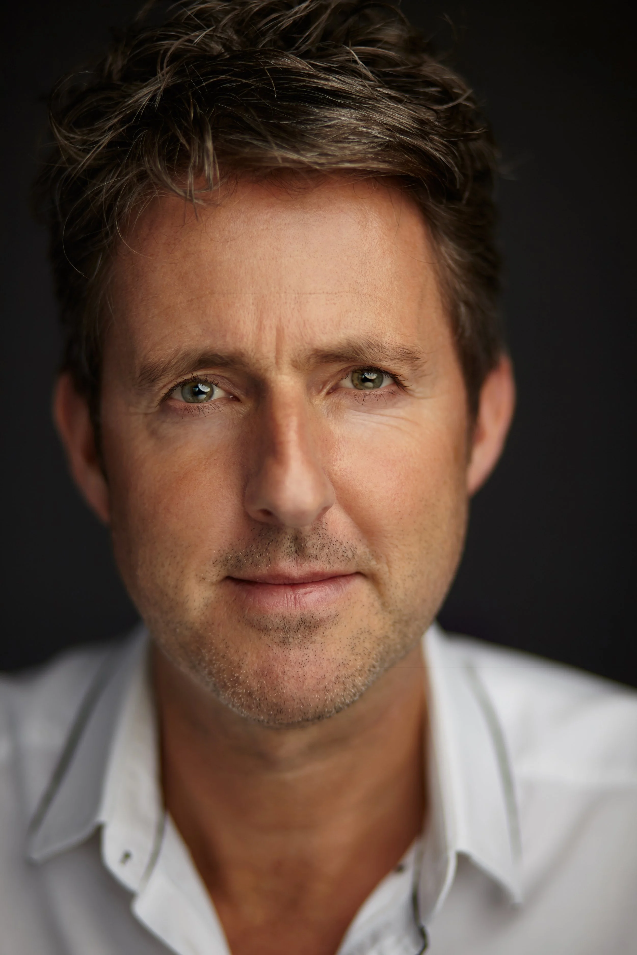 Close-up of a middle-aged man with light brown hair, green eyes, and light stubble, wearing a white shirt with dark piping, against a black background.