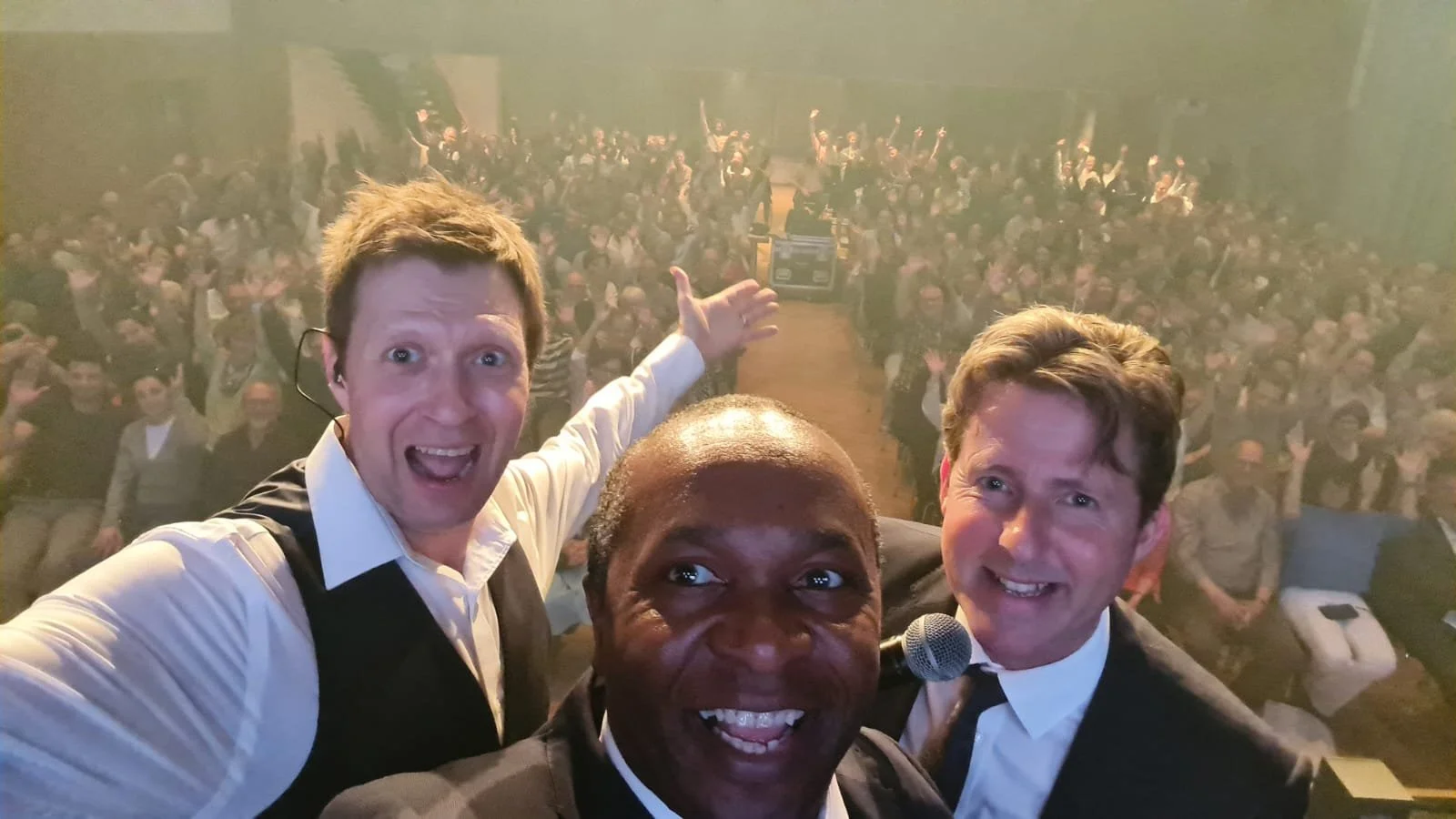 Three men taking a selfie on stage in front of a large, cheering crowd at an event or conference