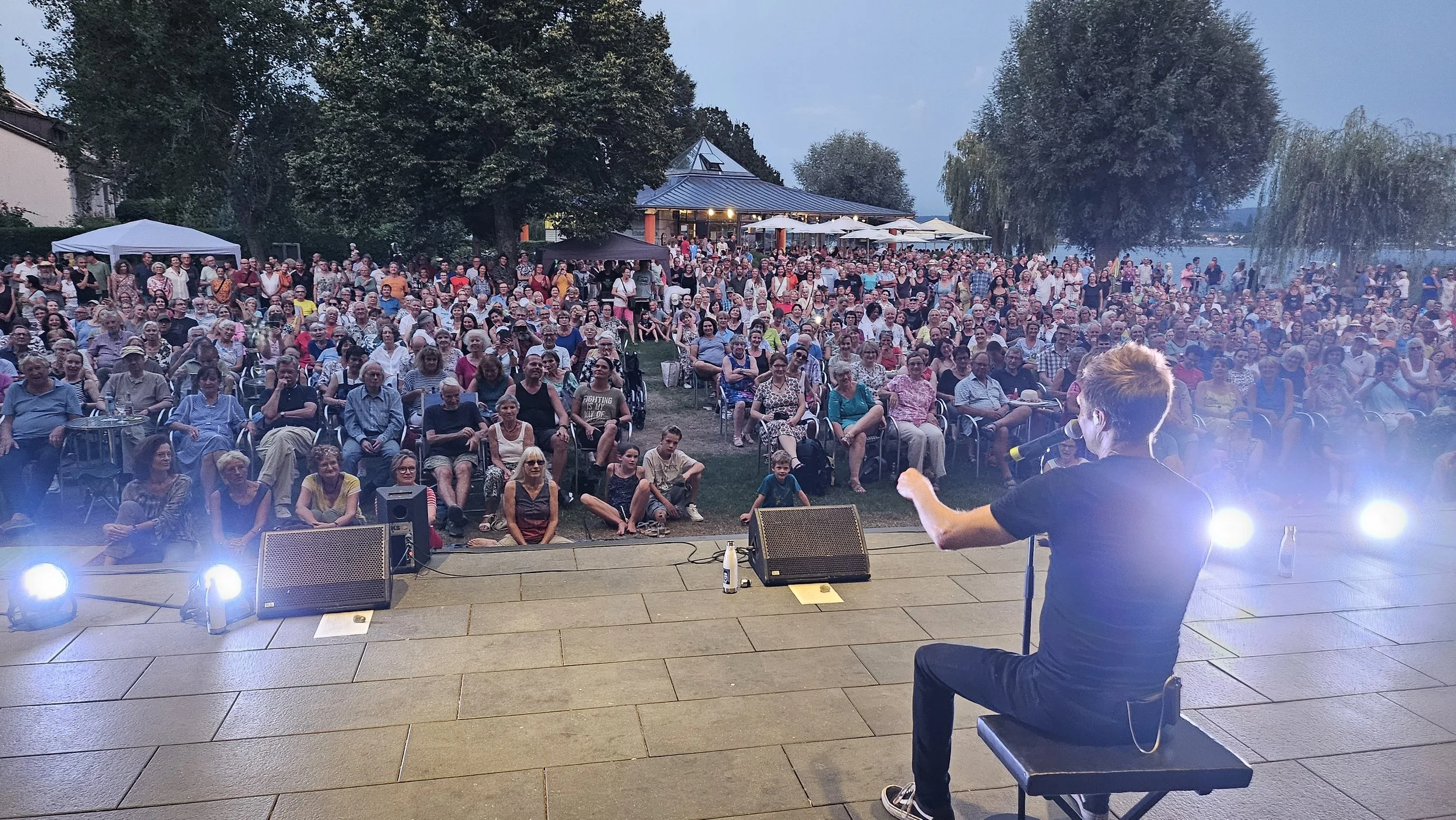 A performer on stage in front of a large audience outdoors, with trees and a building in the background during evening.