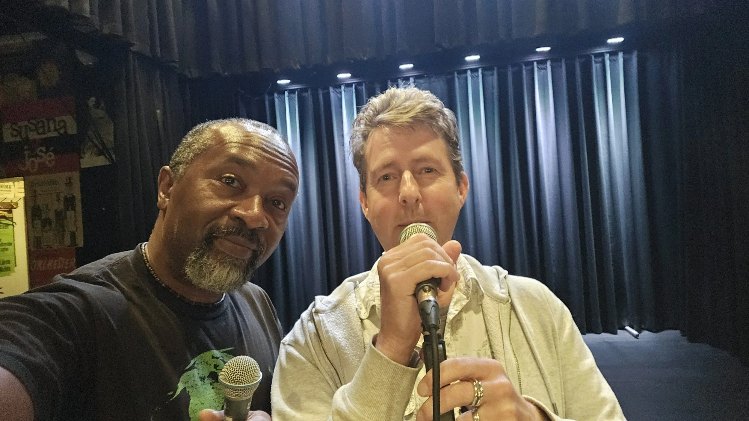 Two men on stage, one holding a microphone and singing, the other taking a selfie with the stage in the background and blue curtains.