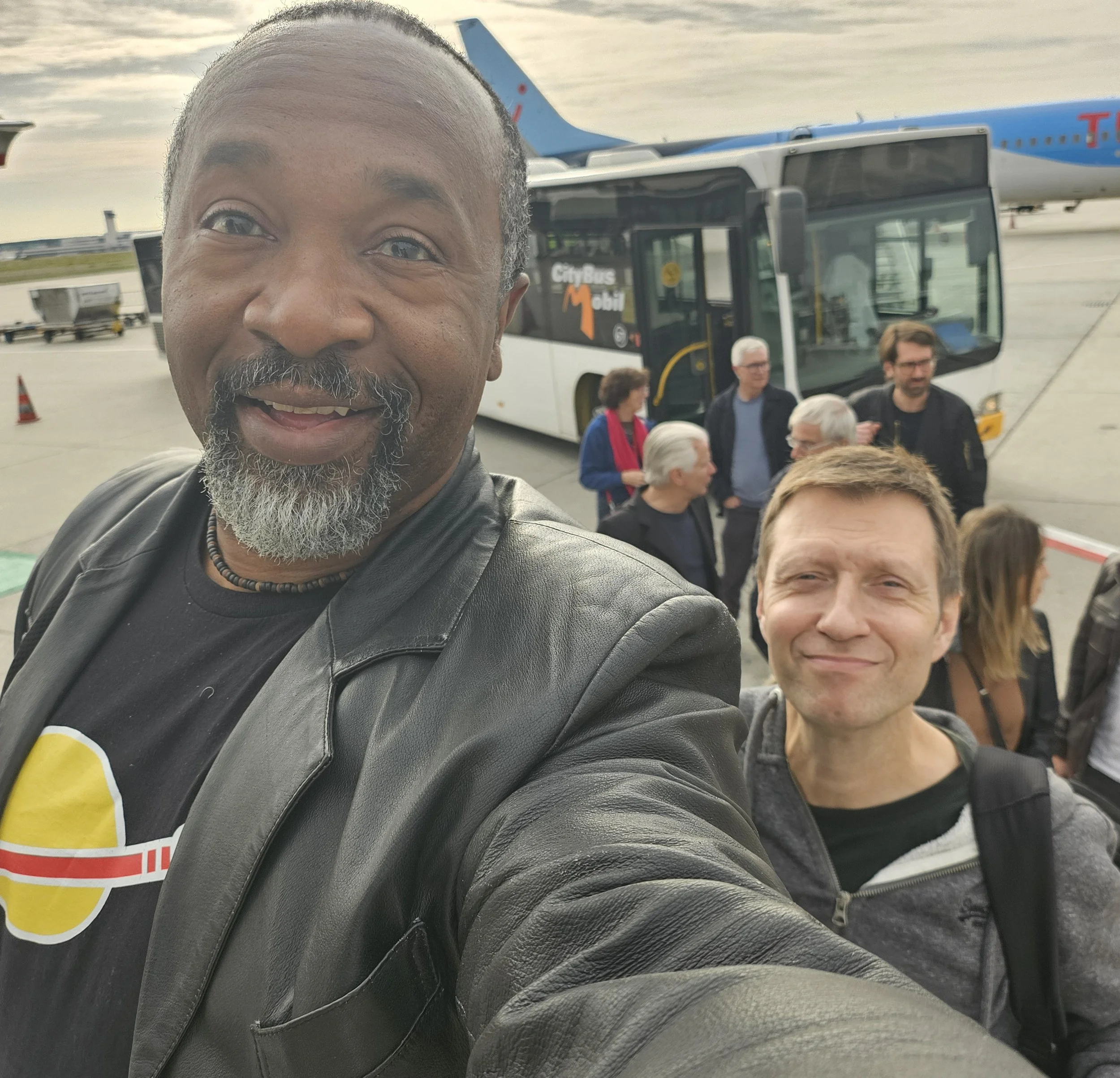 Close-up of a man smiling in front of an airport TUI airplane and a city bus with a small crowd of people in the background.