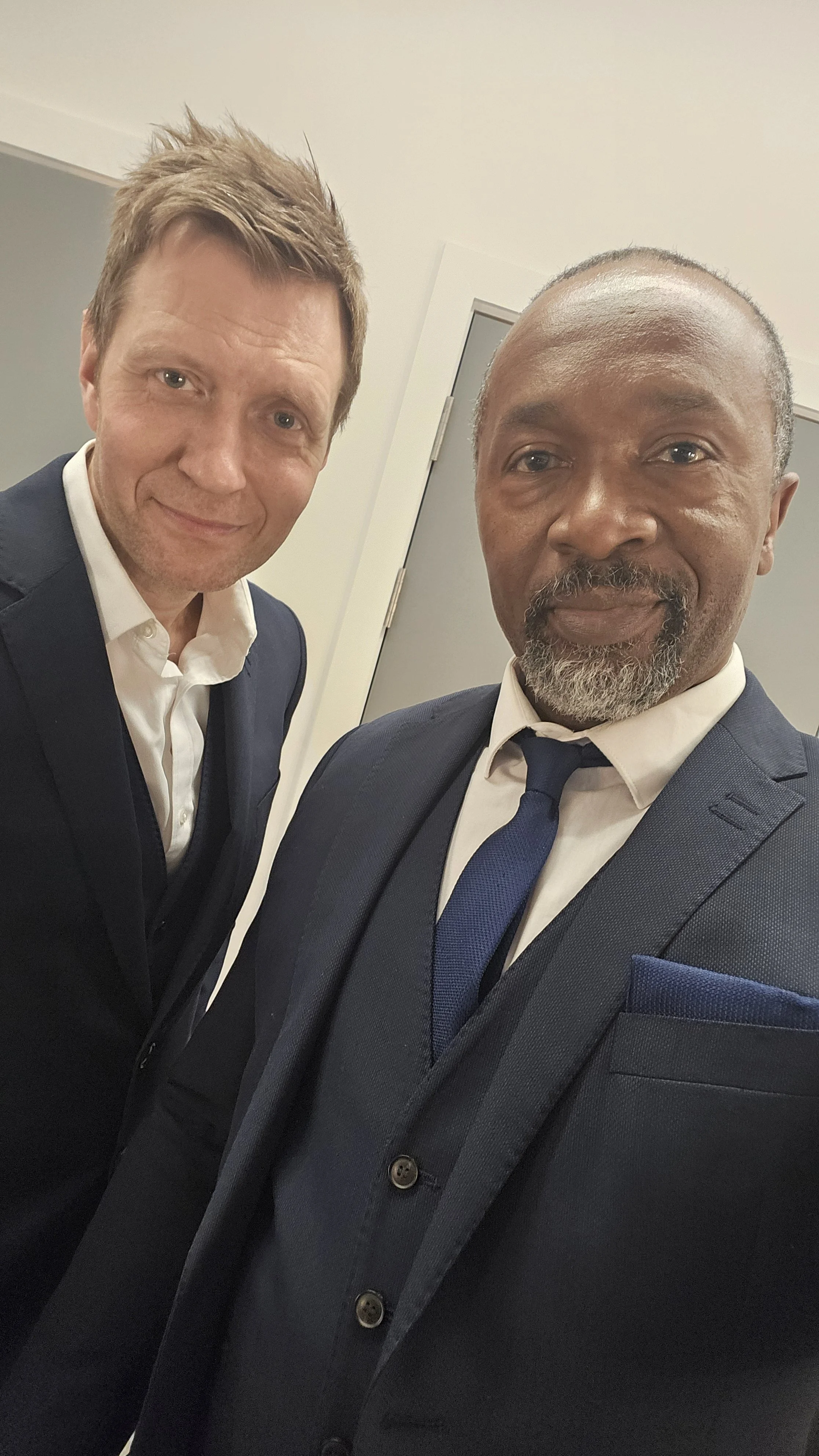 Two men in suits taking a selfie in an indoor setting.