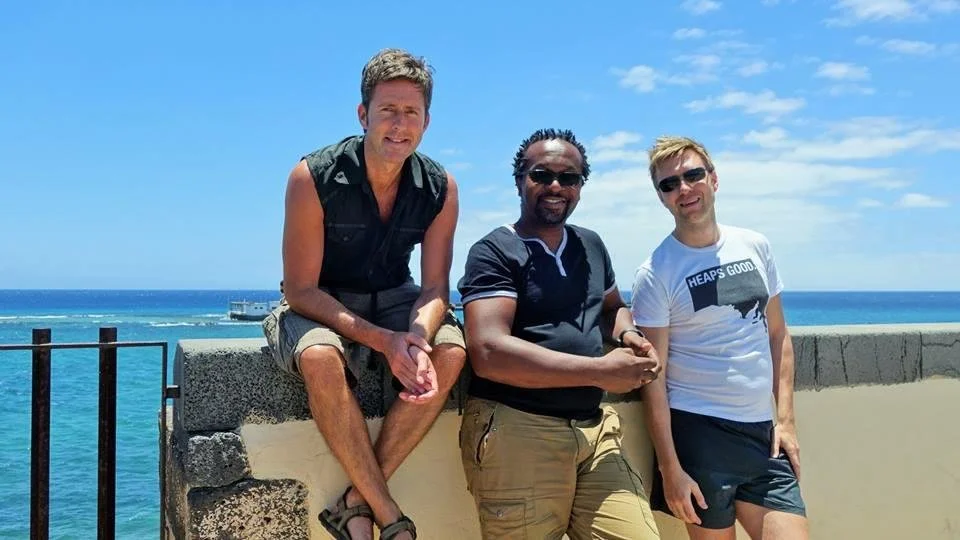 Three men stand together on a seaside promenade with blue sky and ocean in the background.