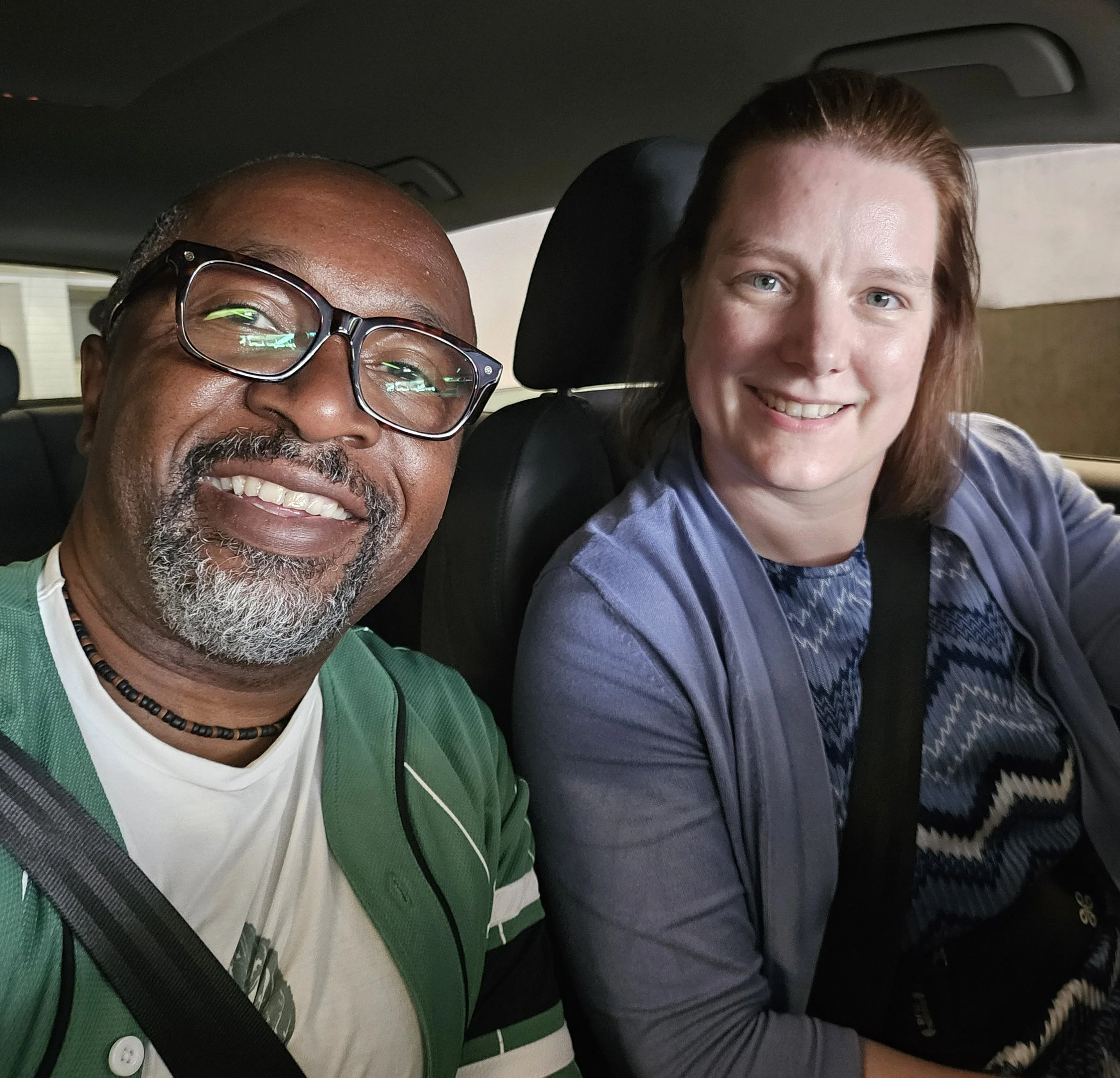 Close-up of a smiling middle-aged man with glasses and a beard, and a young woman with red hair and blue eyes, sitting inside a car.