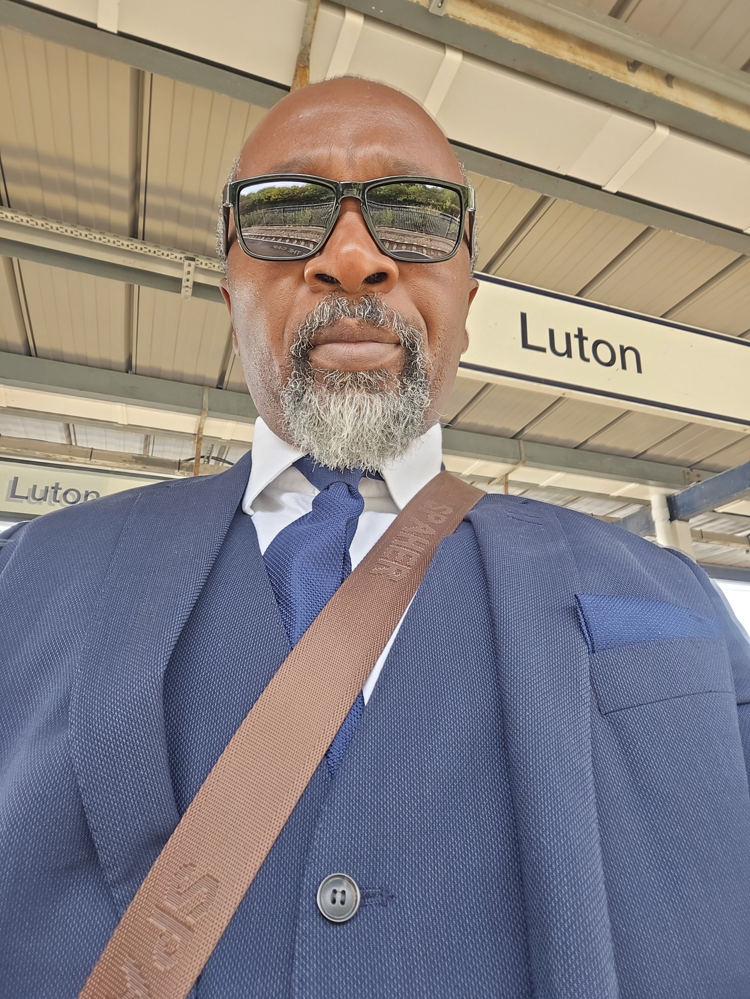 Close-up of a middle-aged man wearing sunglasses, a navy blue suit, white shirt, and navy tie at a train station platform with a sign that says 'Luton' in the background.