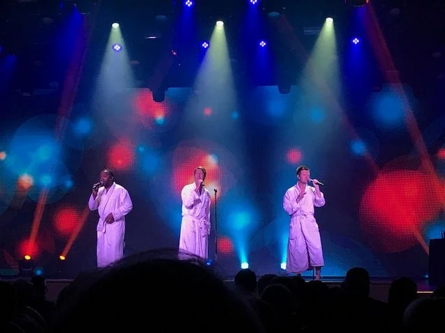Three male singers in white robes performing on stage with colorful stage lights and a digital backdrop.