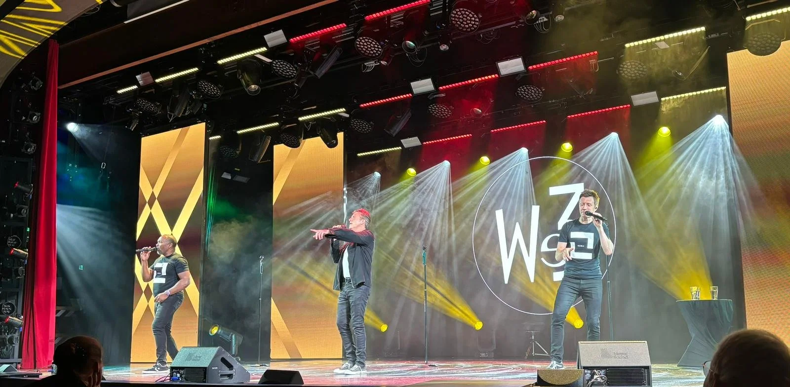 Three men are performing on stage with microphones at a concert or conference. The stage is decorated with colorful lights and large screens displaying the Woz logo. There are fog effects and a table with glasses of water on the right side.