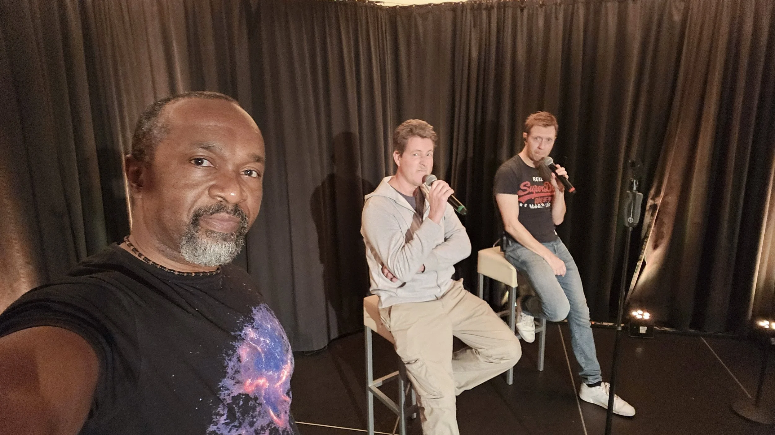 Four men are sitting on stools on a stage, all holding microphones. The man closest to the camera is taking a selfie, with a dark curtain background behind them.