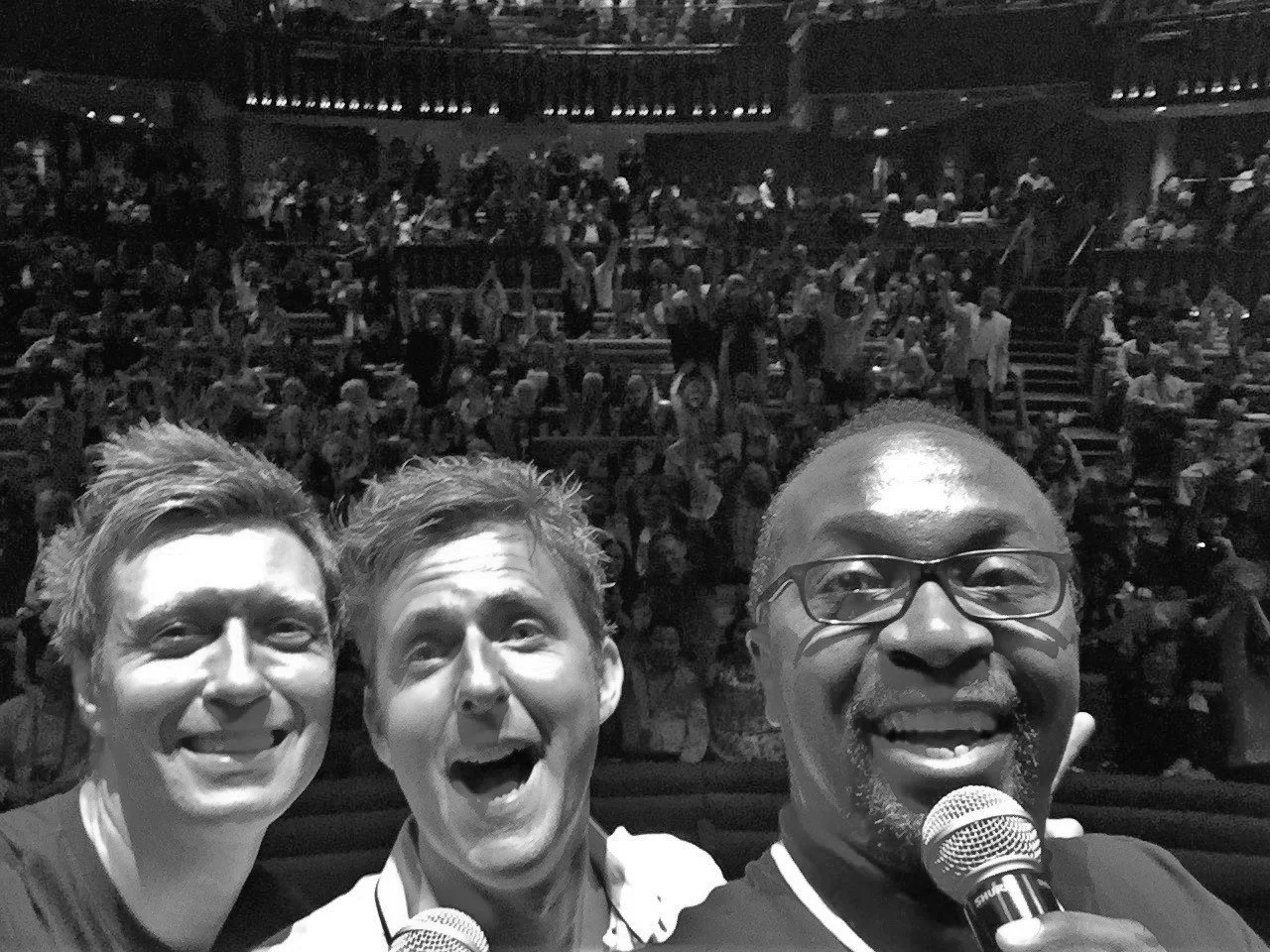 Three men taking a selfie on stage with a large audience in the background, all smiling and holding microphones.