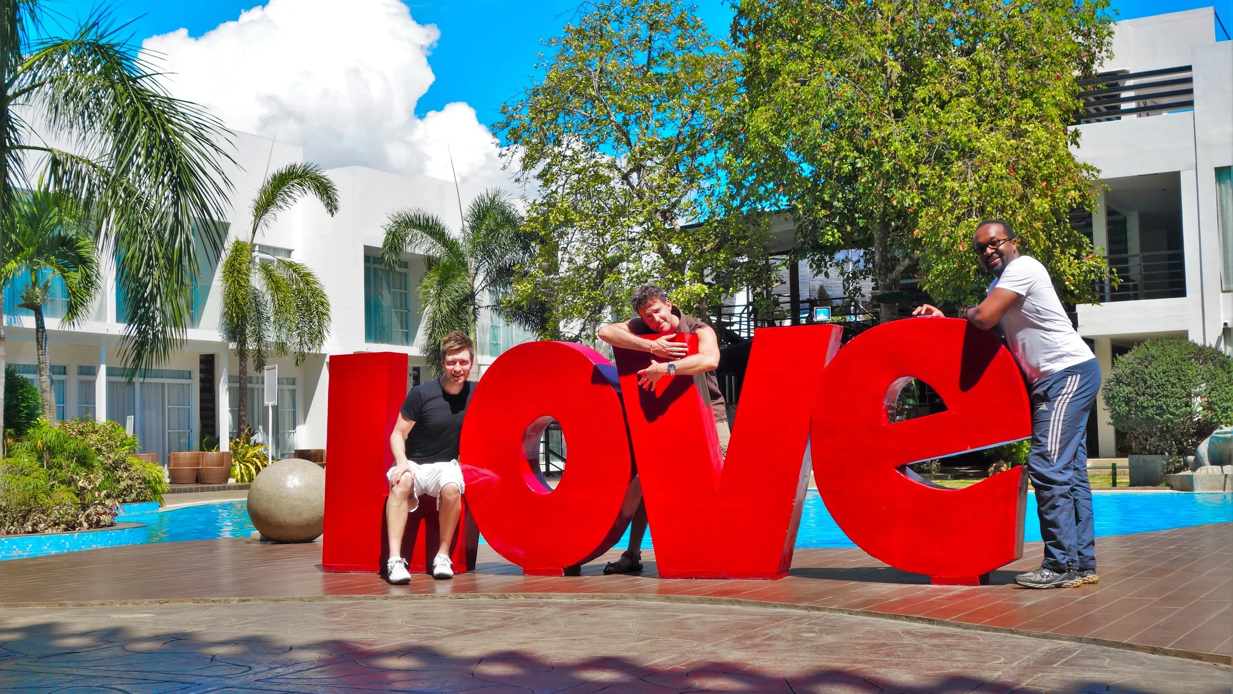 Three men posing with a large red 'LOVE' sculpture near a pool, surrounded by palm trees and modern white buildings, on a sunny day.