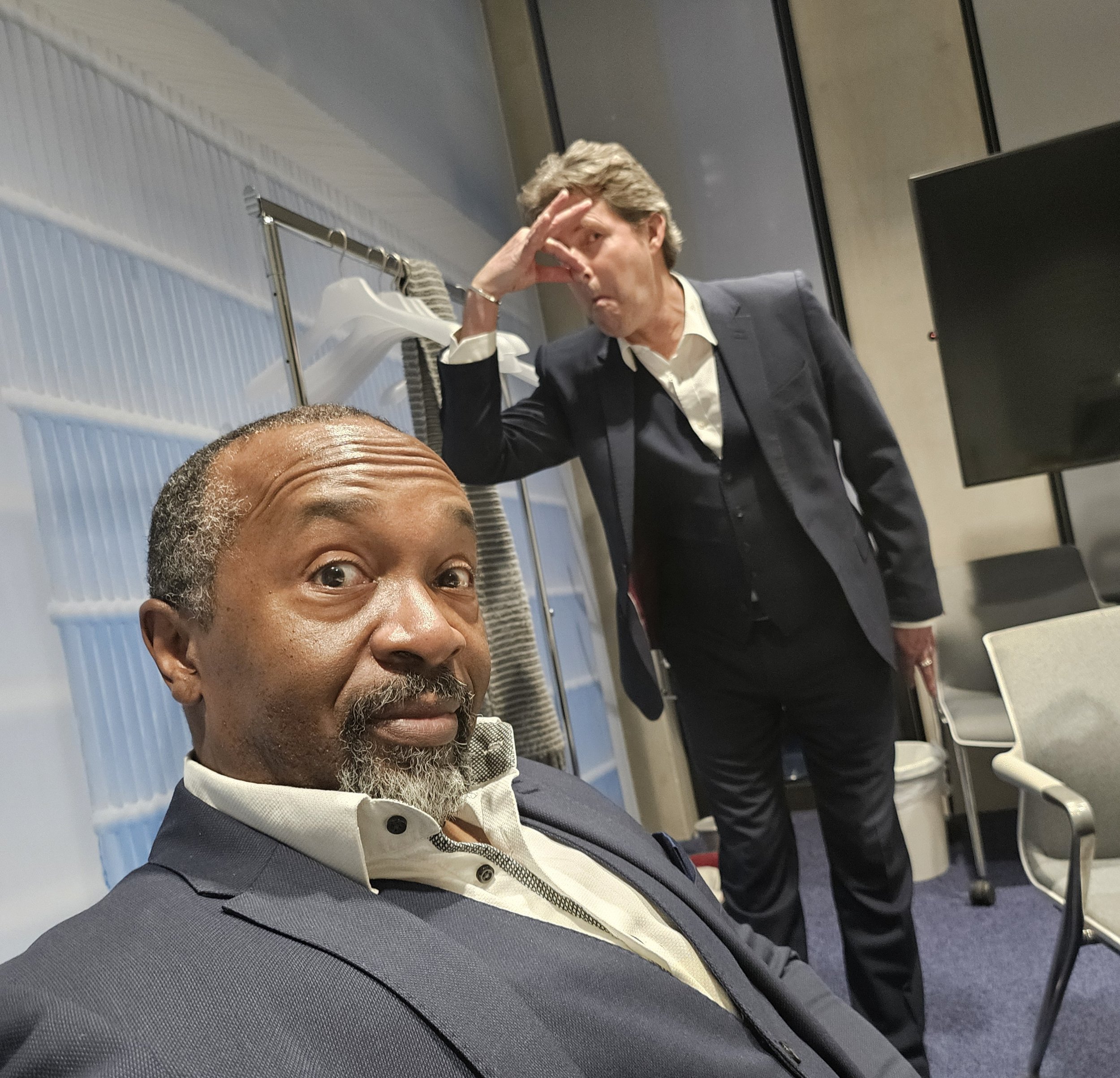 A middle-aged man with a beard and short hair, dressed in a dark suit and white shirt, is sitting on a seat, looking at the camera. There is a man standing in the background, wearing a dark suit and white shirt, with his hand on his forehead, appeari