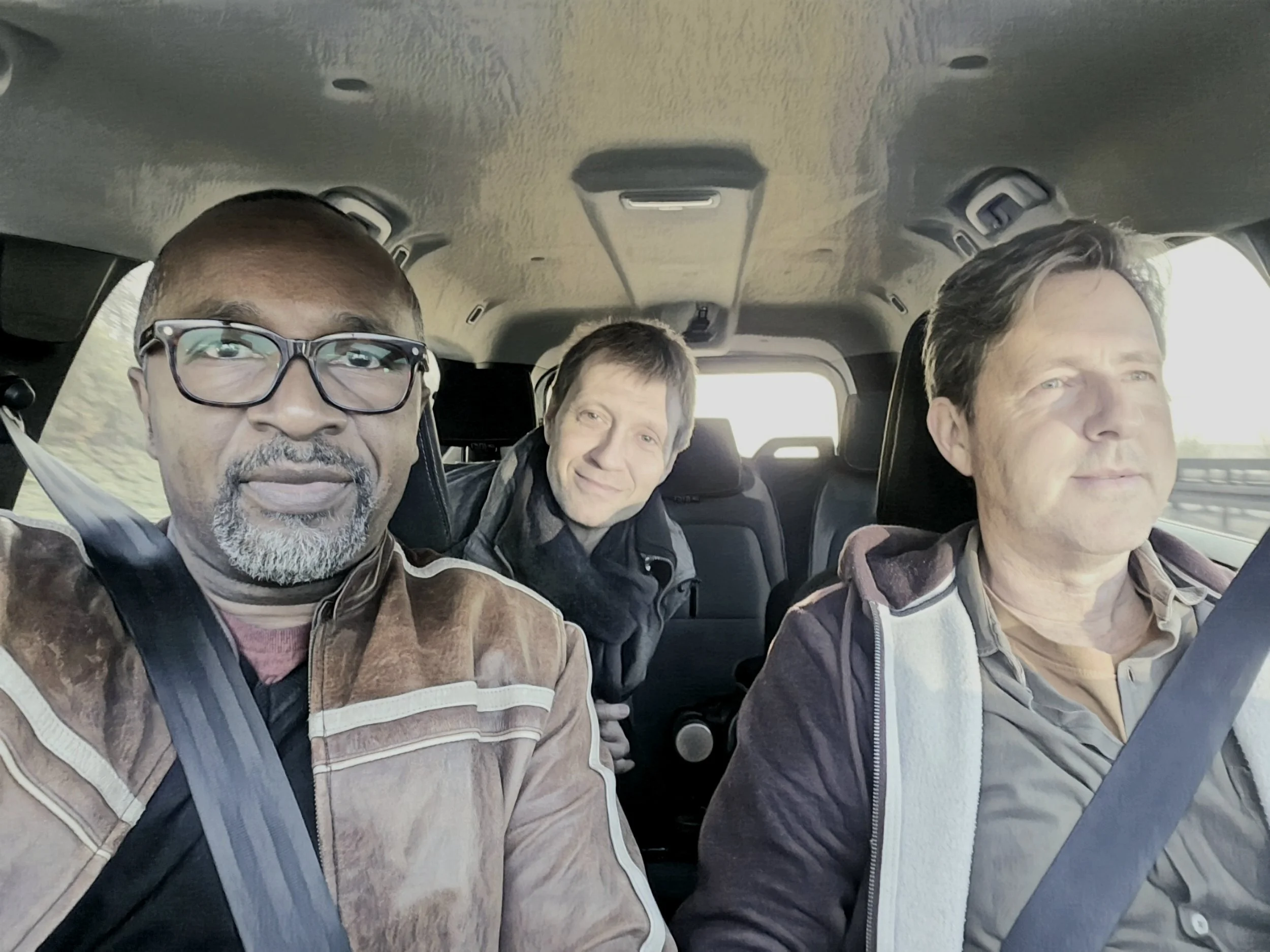 Three men riding in a car on a road, with one man in the driver seat and two men in passenger seats, all facing forward.