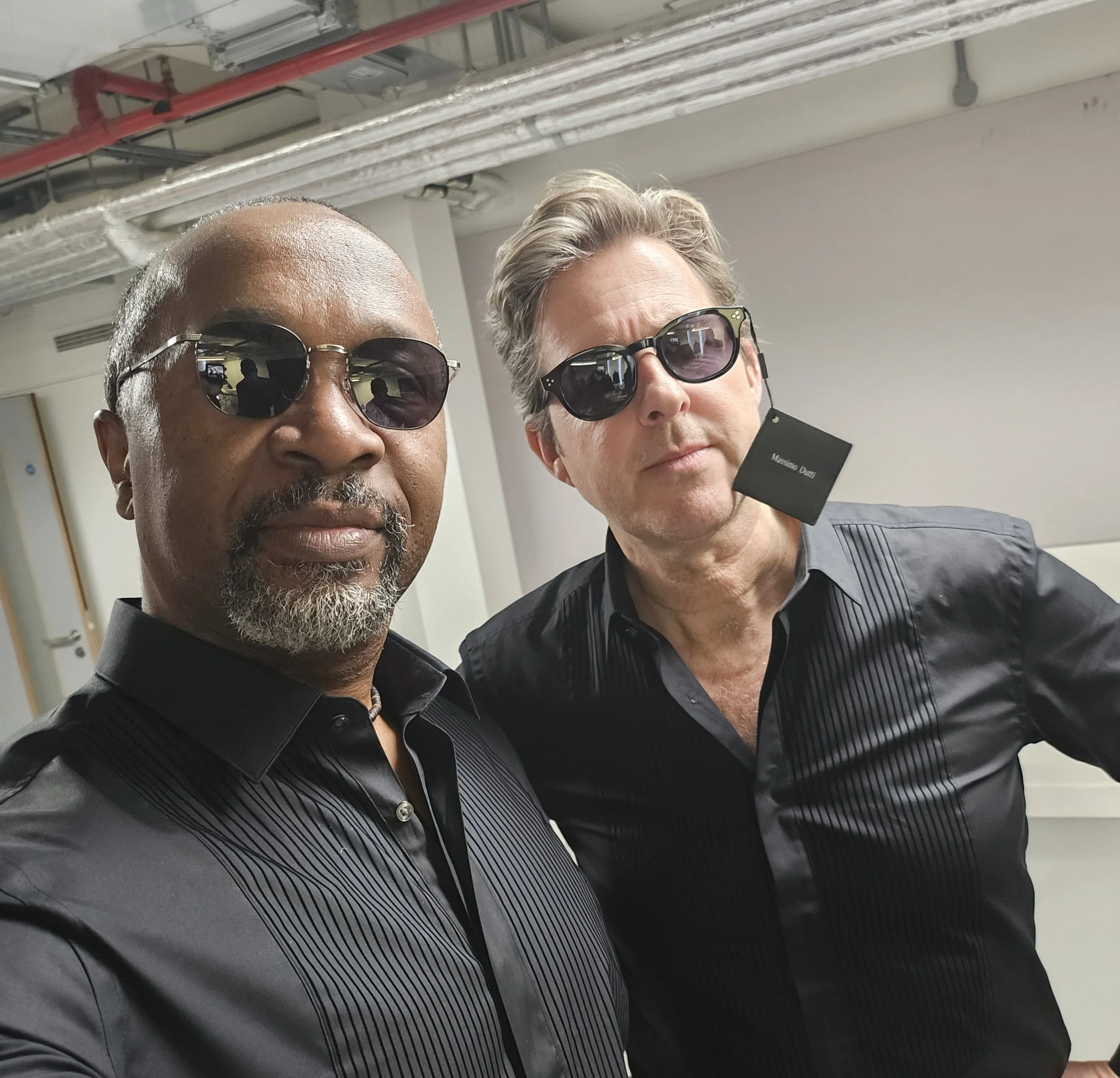Two men wearing dark sunglasses and dark business attire posing for a photo, with an industrial or office background featuring pipes and a tag with the name 'Massimo Dutti'.
