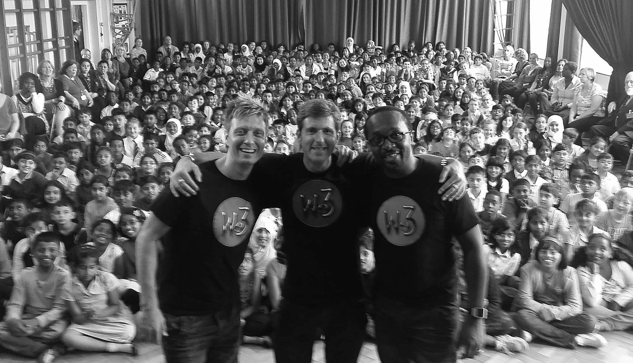 Three men standing in front of a large group of children and adults in a school auditorium, smiling and arms around each other.
