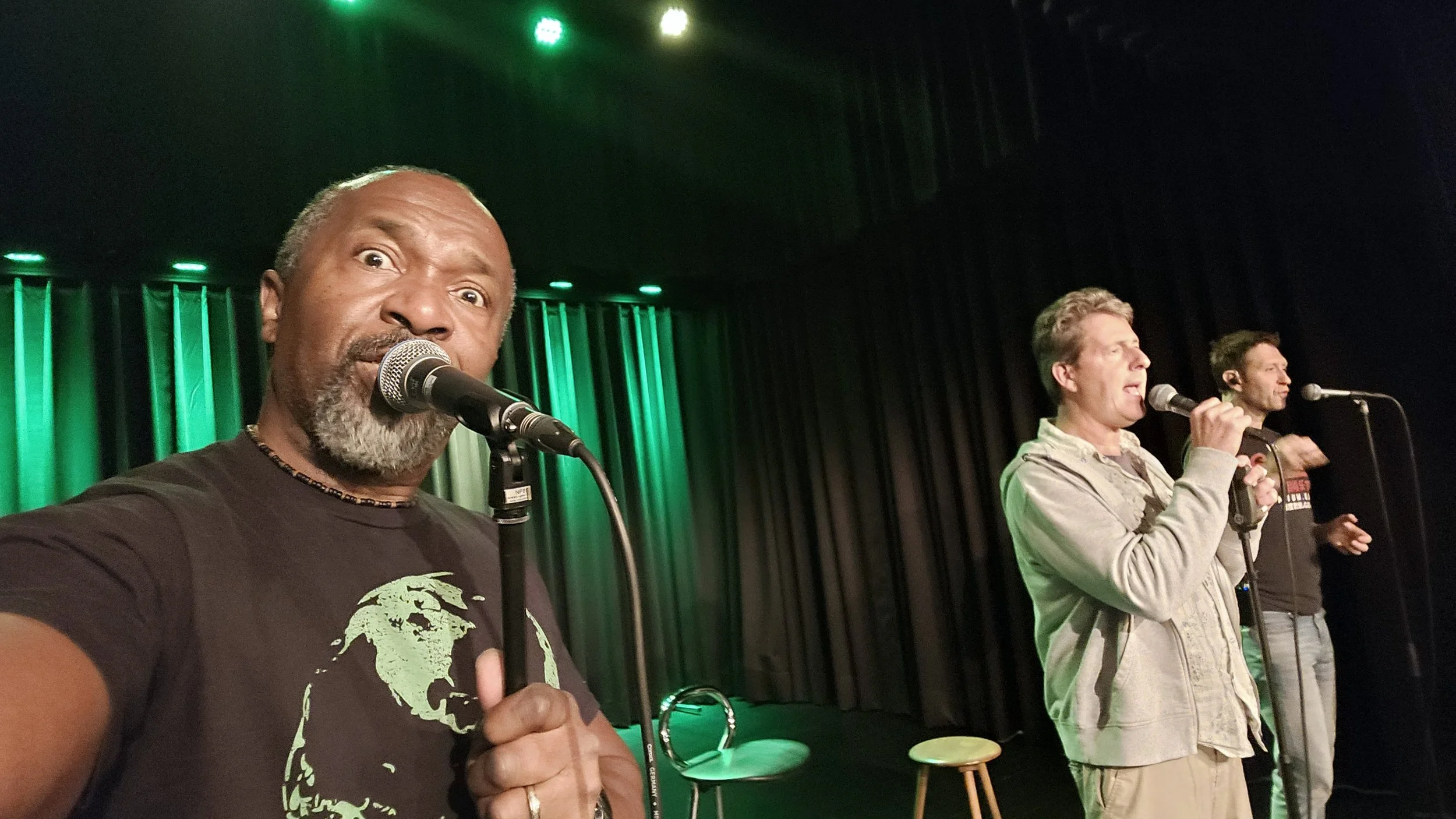 Group of three singerss on stage, with the man in the foreground taking a selfie, all singing into microphones, in front of a dark curtain with green and yellow stage lights.
