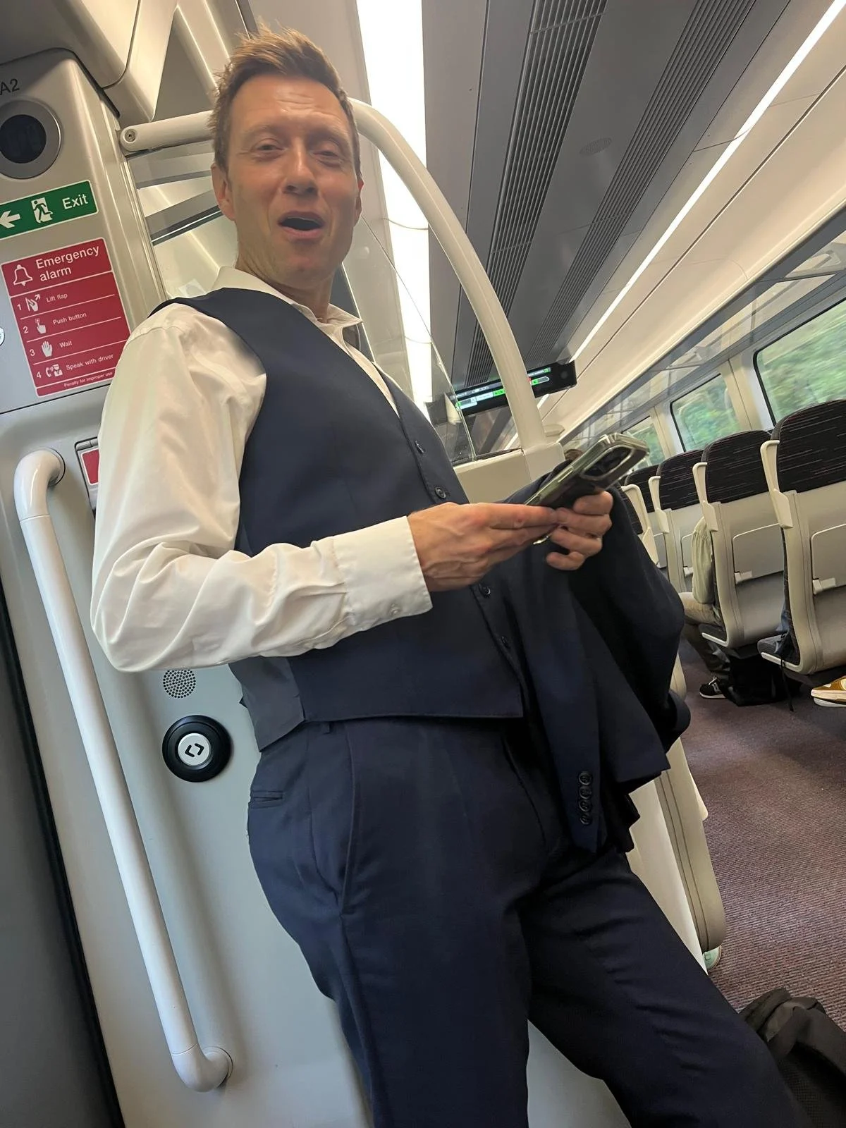 A man in a dress shirt and vest standing inside a train, holding a phone, with train seats and an emergency sign visible in the background.