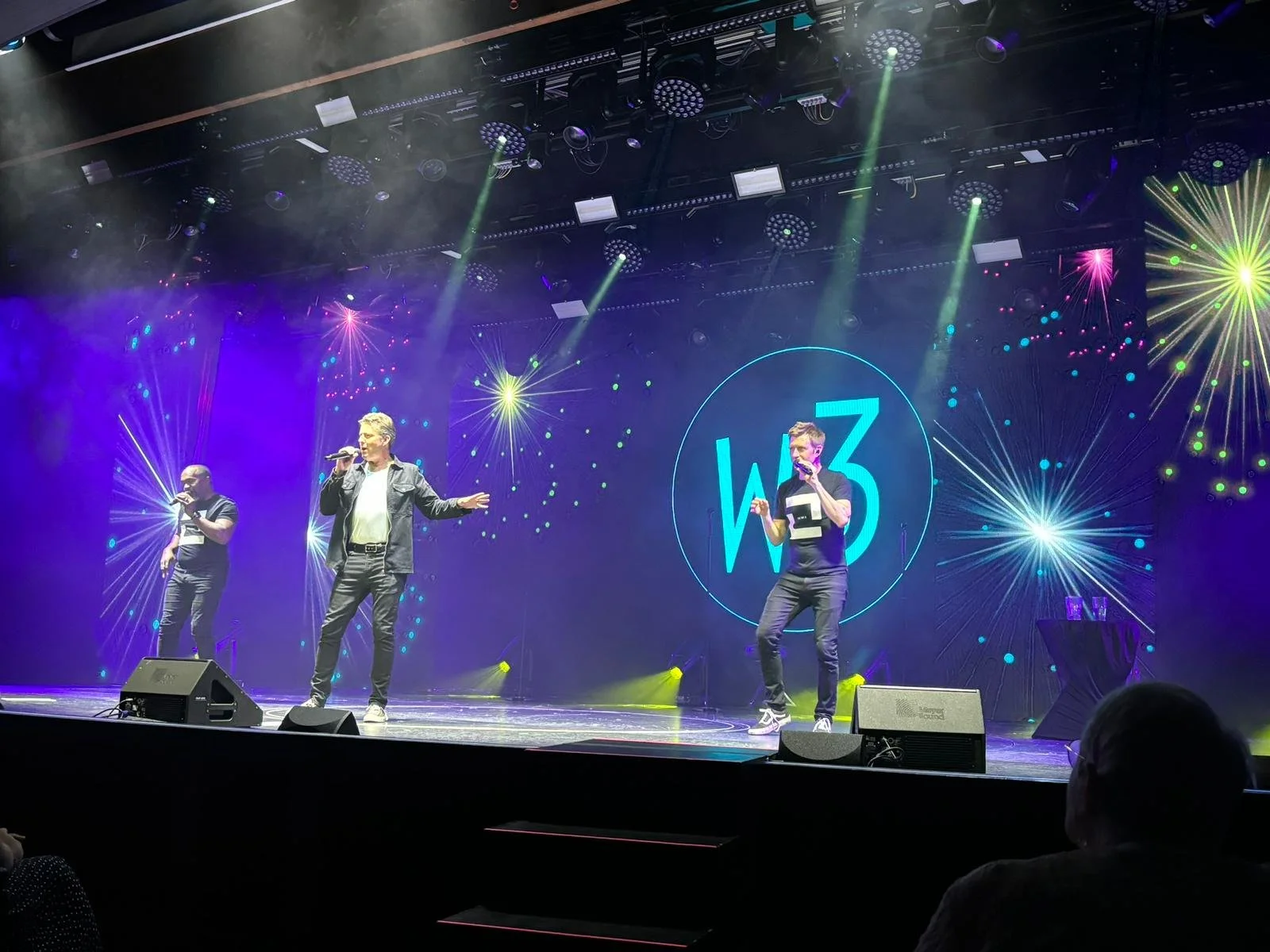 Three men perform on stage under colorful fireworks-like digital visuals. The backdrop displays a circle with the text 'W3'. One man is singing into a microphone, the others are gesturing with microphones in hand. Stage lighting includes beams direct