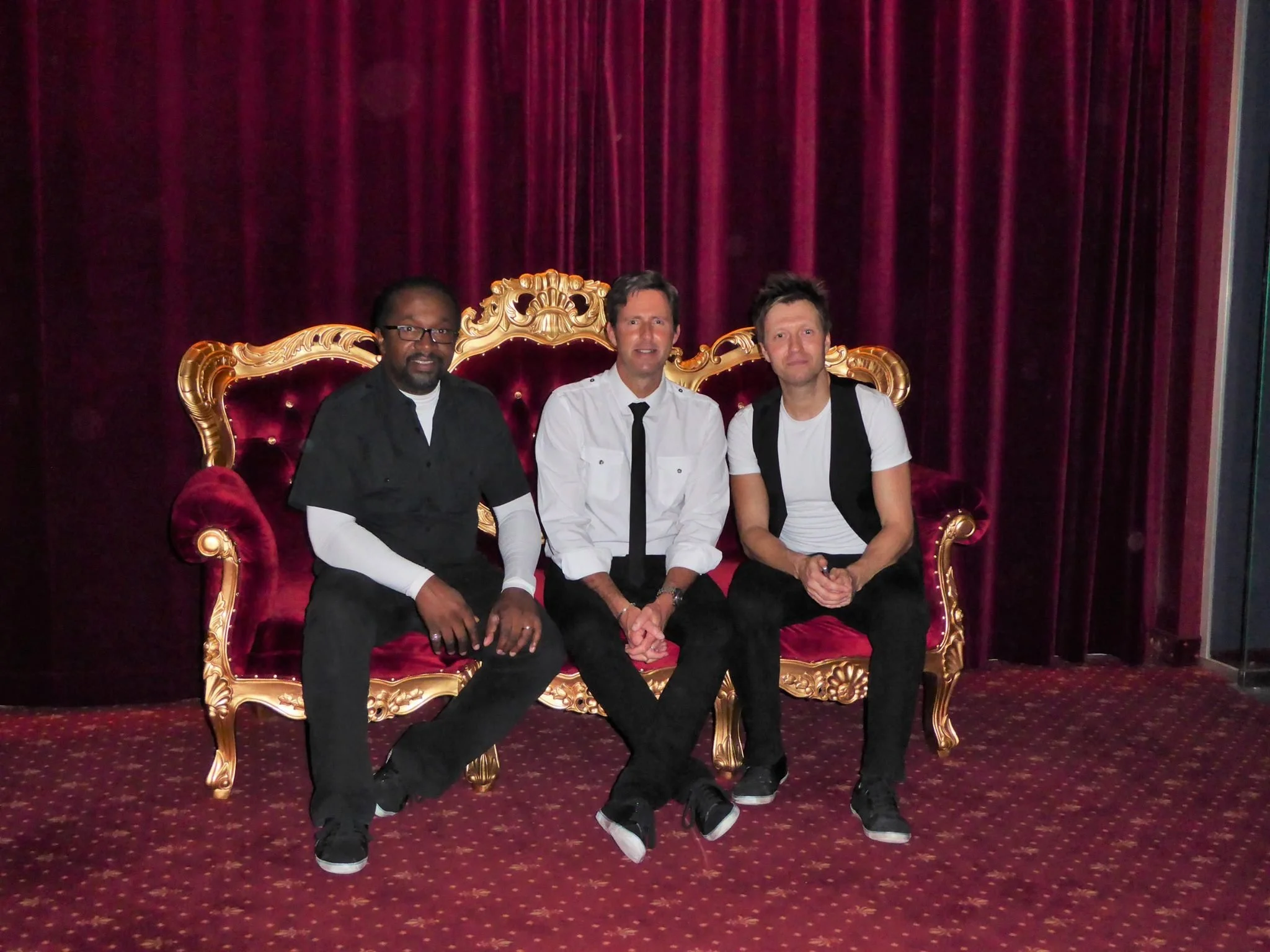 Three men sitting on a luxurious, ornate red velvet and gold royal-style couch in front of a red curtain. The men are wearing casual and semi-formal outfits, with two of them smiling slightly and one appearing to have a neutral expression.