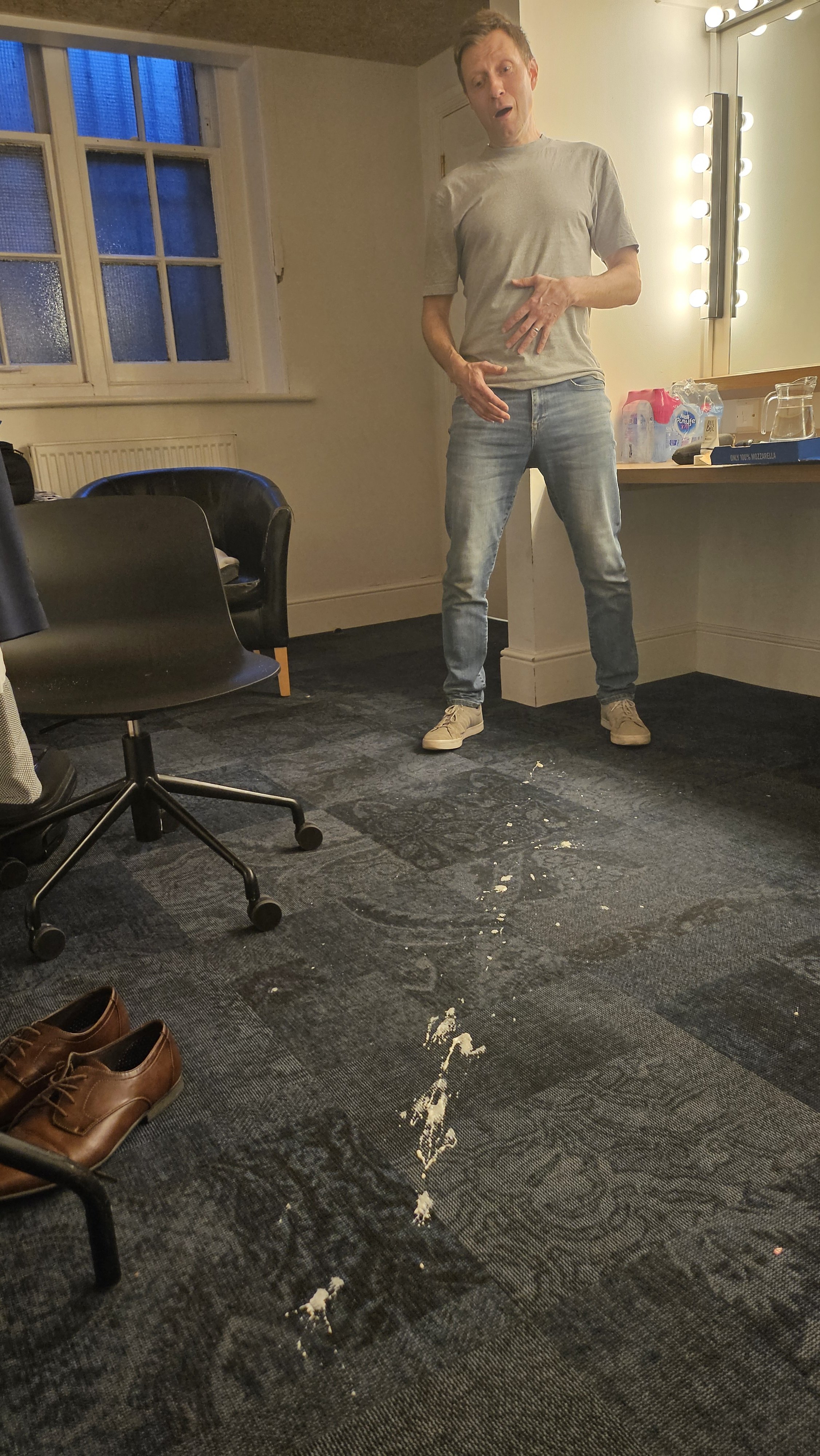 A man standing in a dressing room, reacting with surprise, with some spilled food or beverage on the carpeted floor and shoes in the foreground.