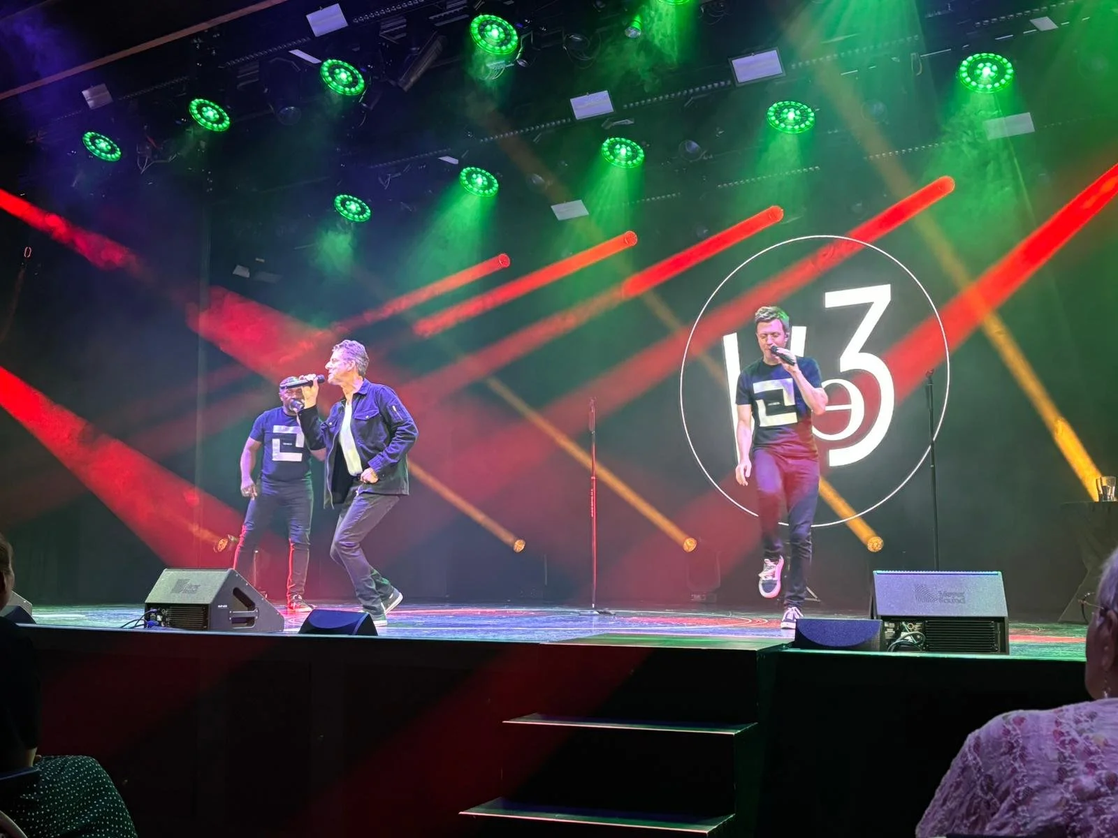 Two male performers singing on a stage with colorful lighting and a large logo in the background.