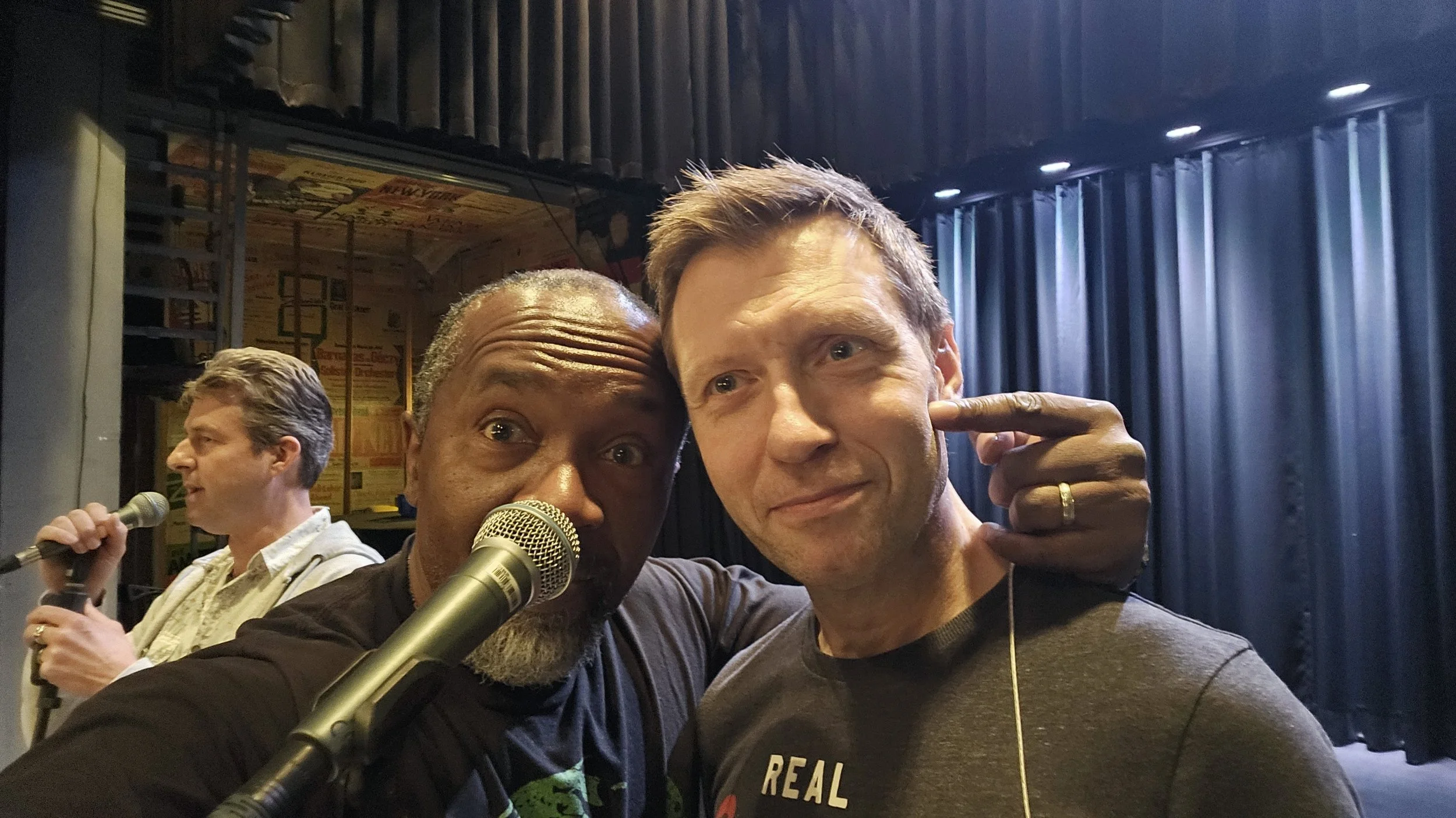 Two men taking a selfie on a stage with microphones; one pointing to his head, with curtains and an additional person speaking into a microphone in the background.