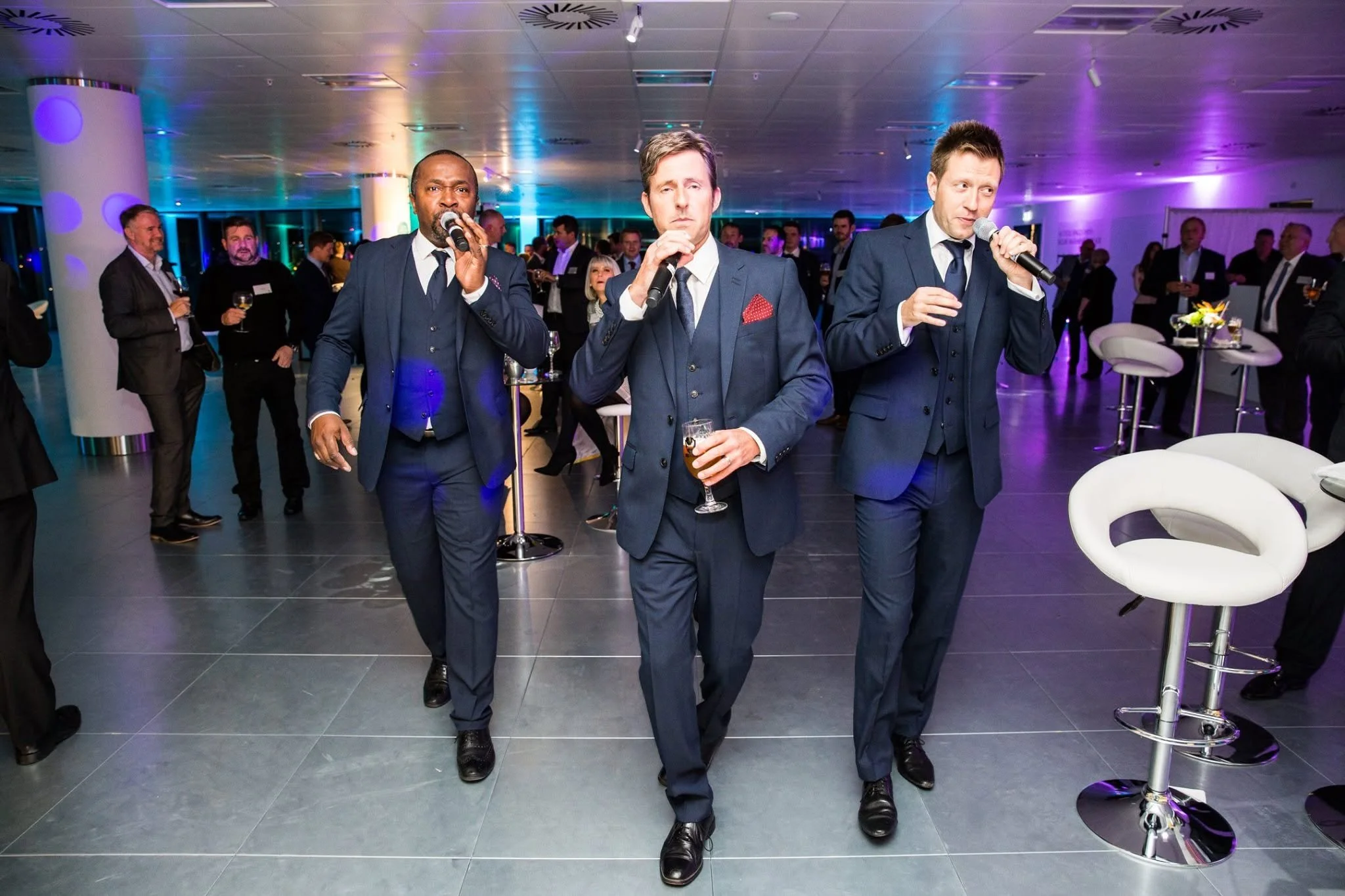 Three men in suits walking through a cocktail party, holding microphones and drinks, with other guests in the background.