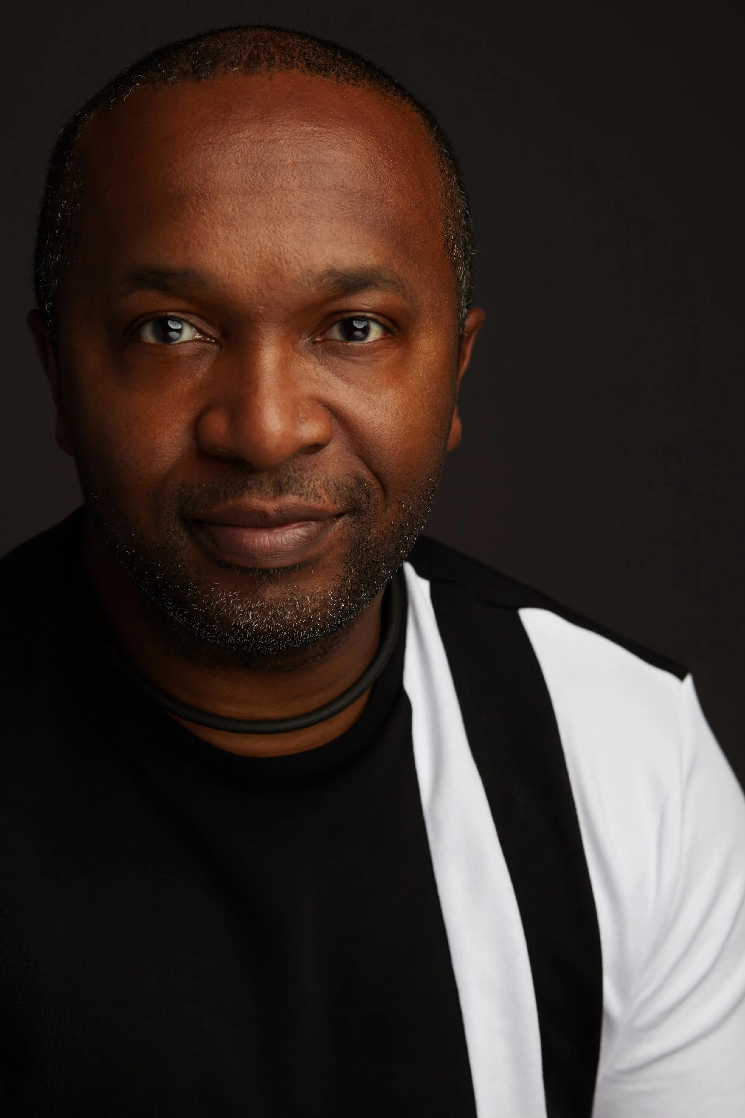 Close-up portrait of a man with a shaved head and gray facial hair, wearing a black and white outfit and a black necklace, against a black background.