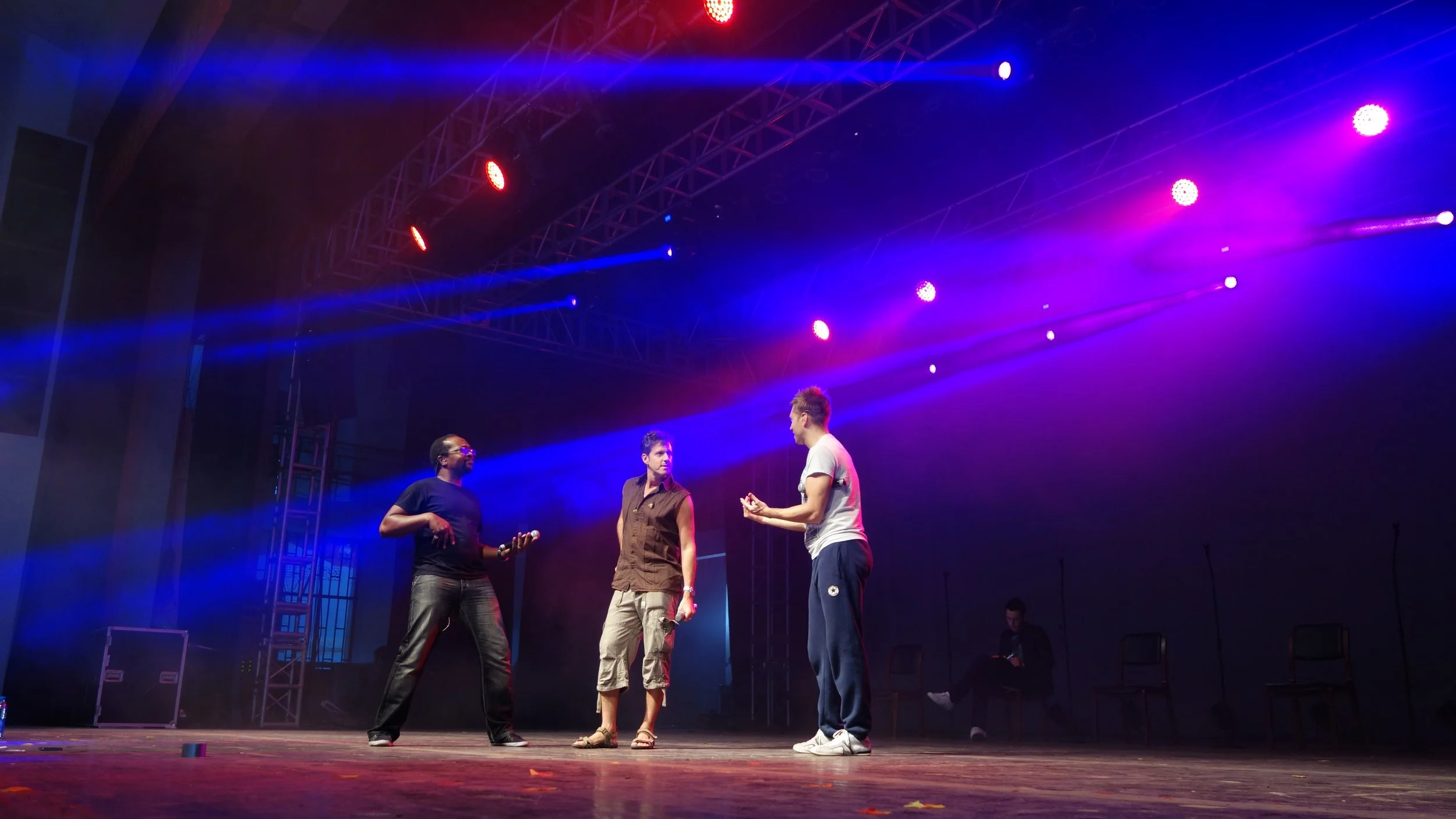 Three men are standing on a stage under colorful red, blue, and purple stage lights. They are engaged in conversation, with one man wearing a white t-shirt, another in a vest, and the third in a black t-shirt. There are chairs and equipment visible i