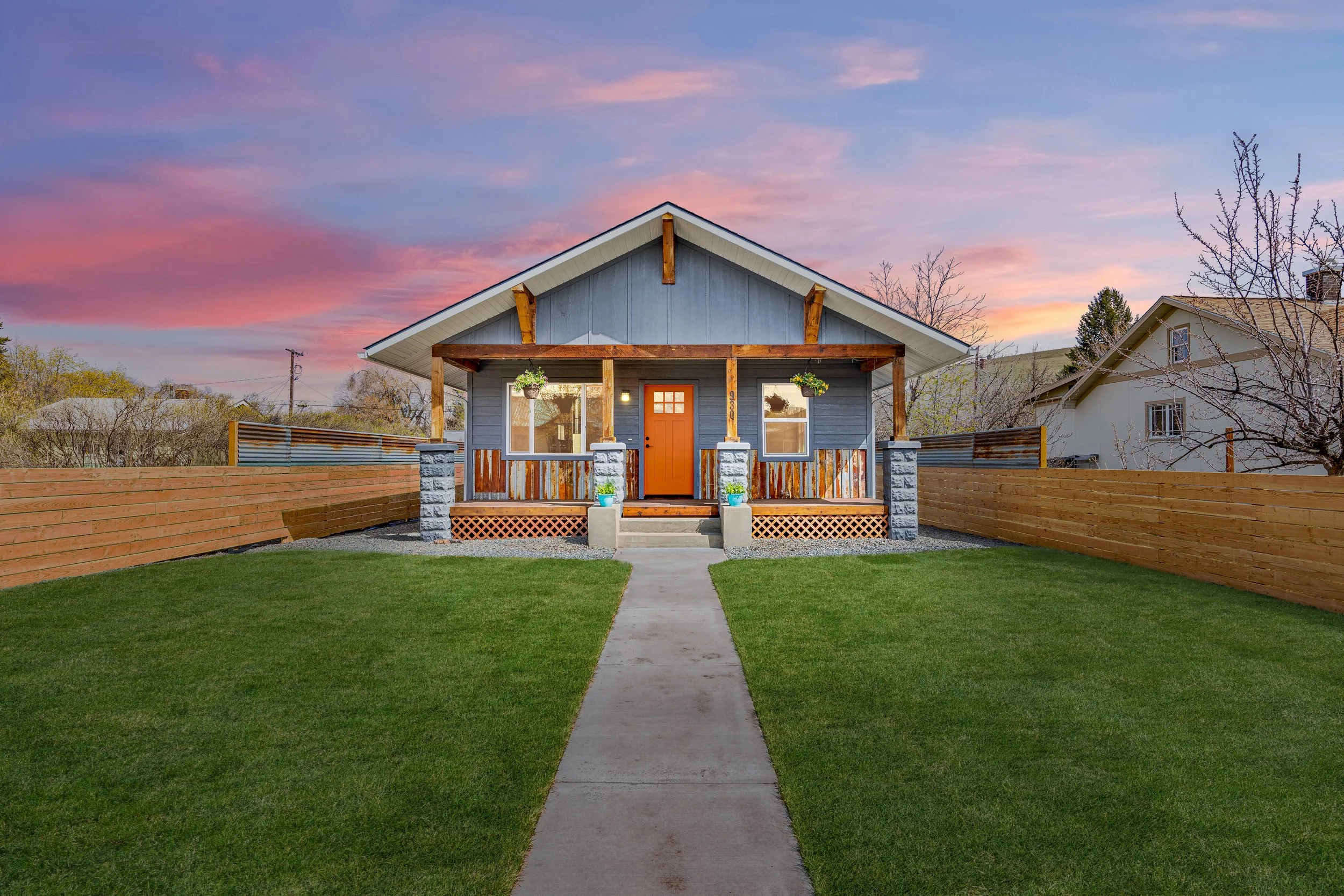 Front view of a small blue house with a porch, orange door, two windows, hanging flower pots, and a wooden fence, under a colorful sunset sky.