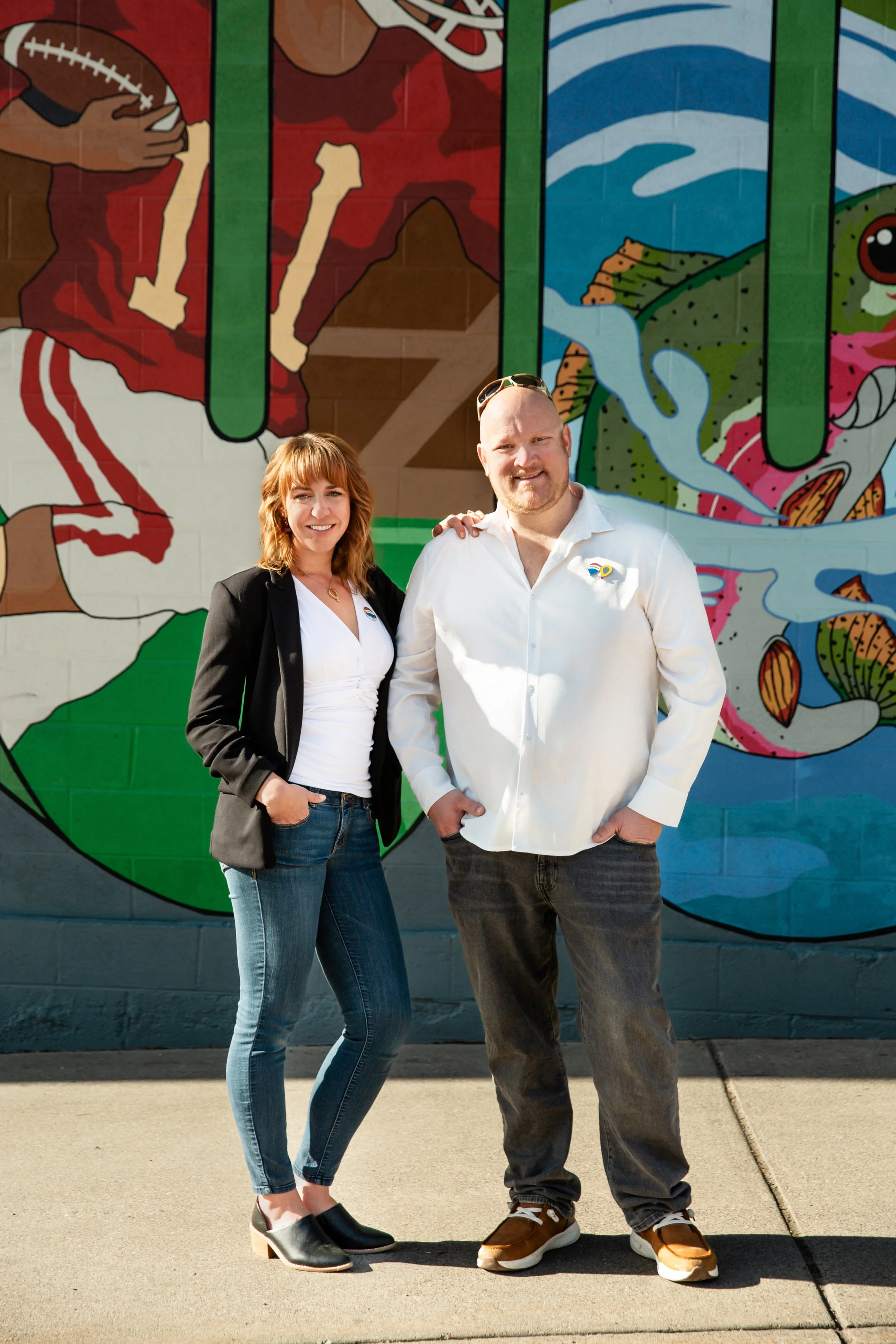 A woman and a man standing in front of a colorful mural, smiling at the camera. The woman has red hair, is wearing a black blazer, white top, and jeans. The man is wearing a white shirt, gray pants, and sunglasses on his head.