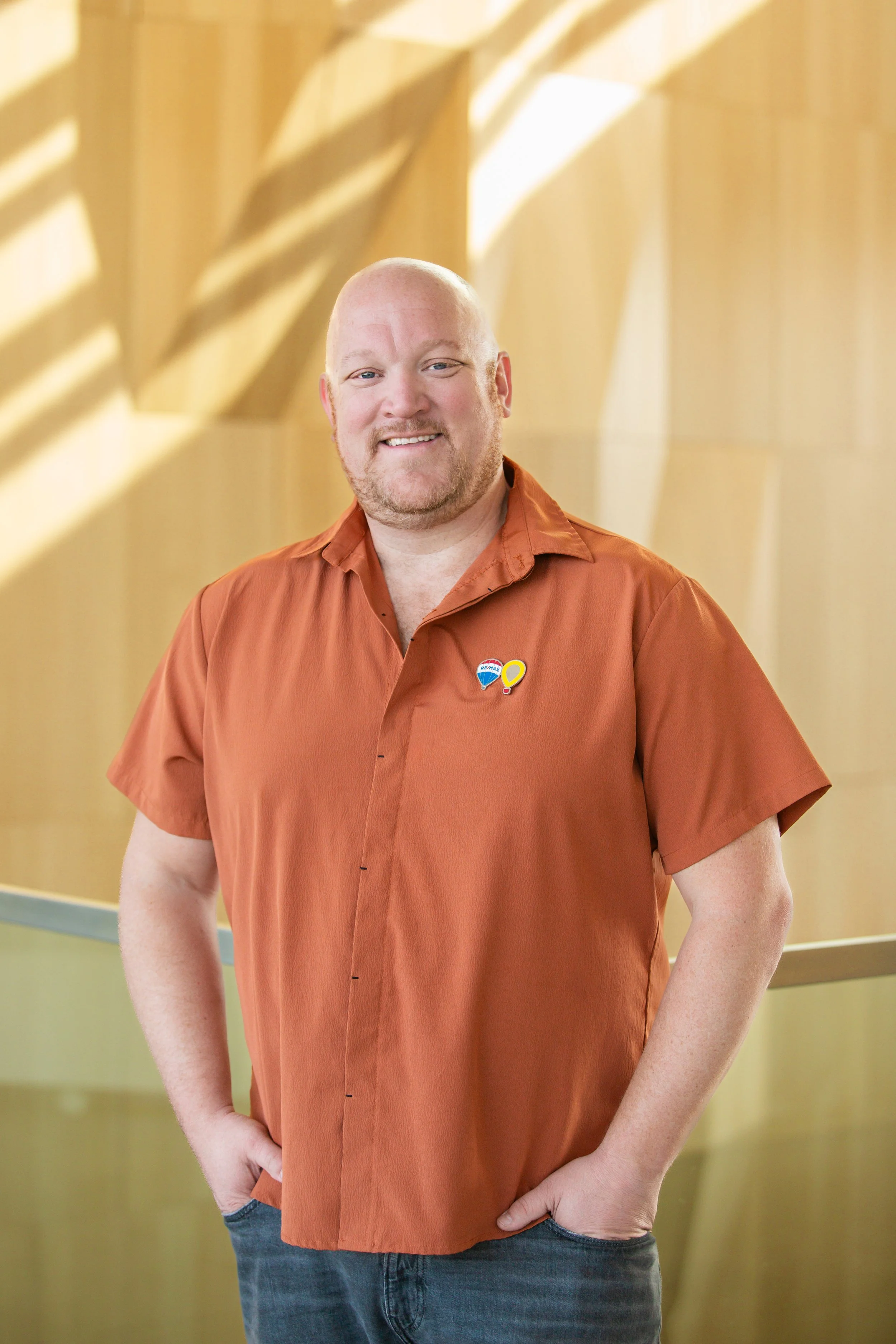 A man with a bald head, goatee, wearing an orange short-sleeved shirt with a pin on the chest standing in front of a yellow wall with shadows.