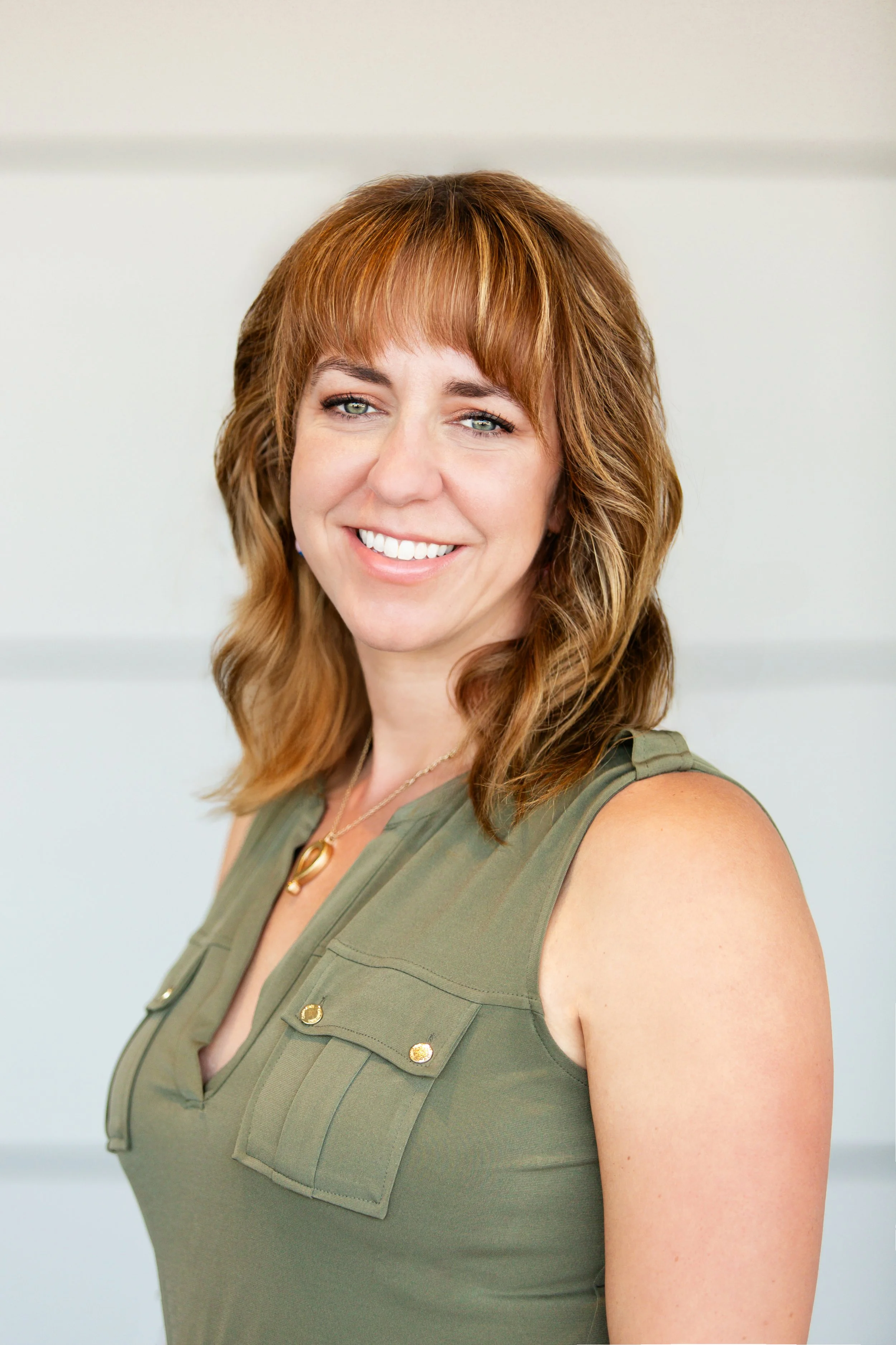 A woman with shoulder-length auburn hair, wearing a sleeveless green top with button details and a gold necklace, smiling at the camera against a plain light-colored background.