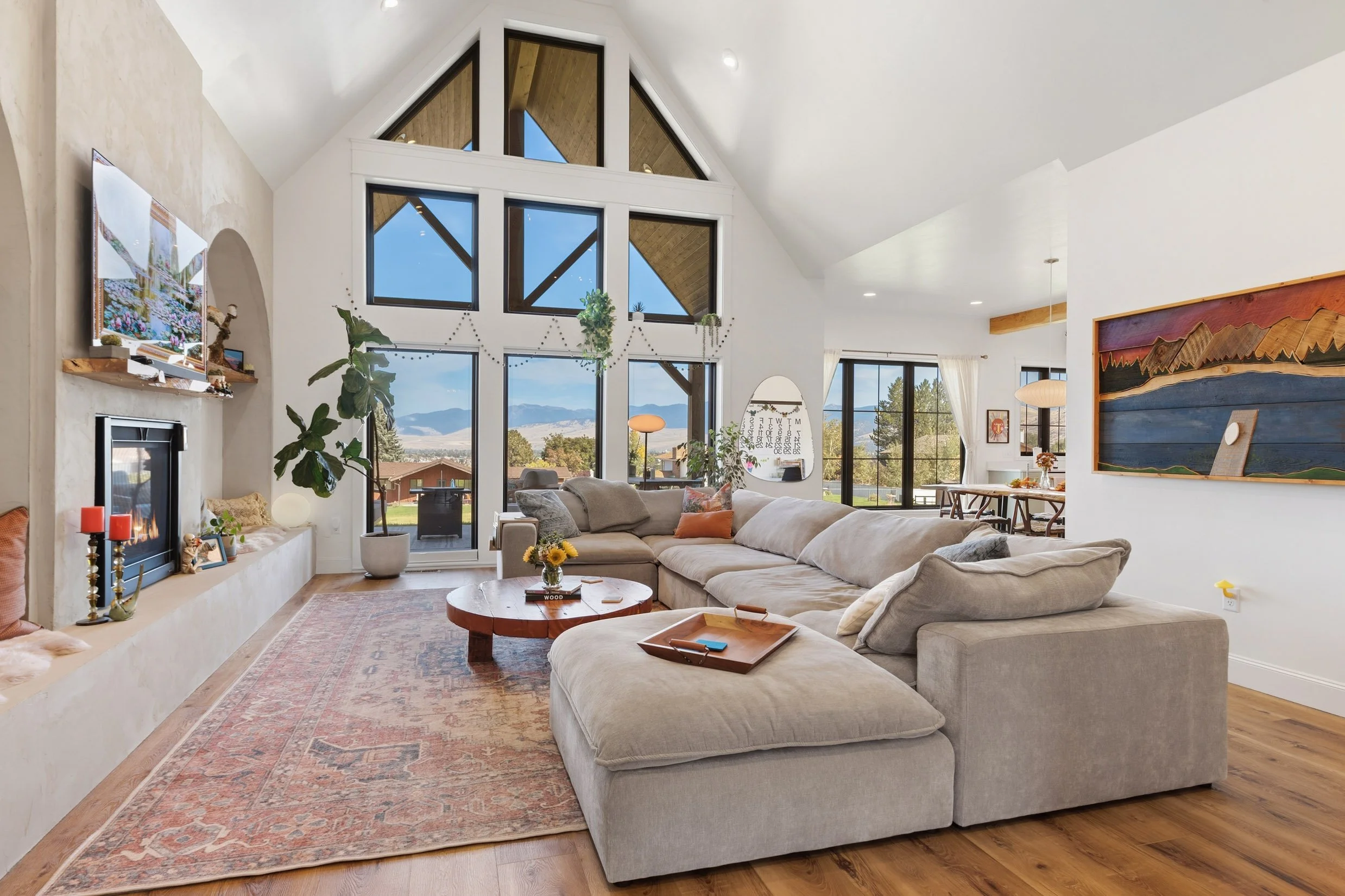 A cozy living room with a large beige sectional sofa decorated with pillows, a round wooden coffee table, a fireplace with a mantel decorated with candles and photos, a staircase, a large window with mountain views, indoor plants, and artwork on the walls.