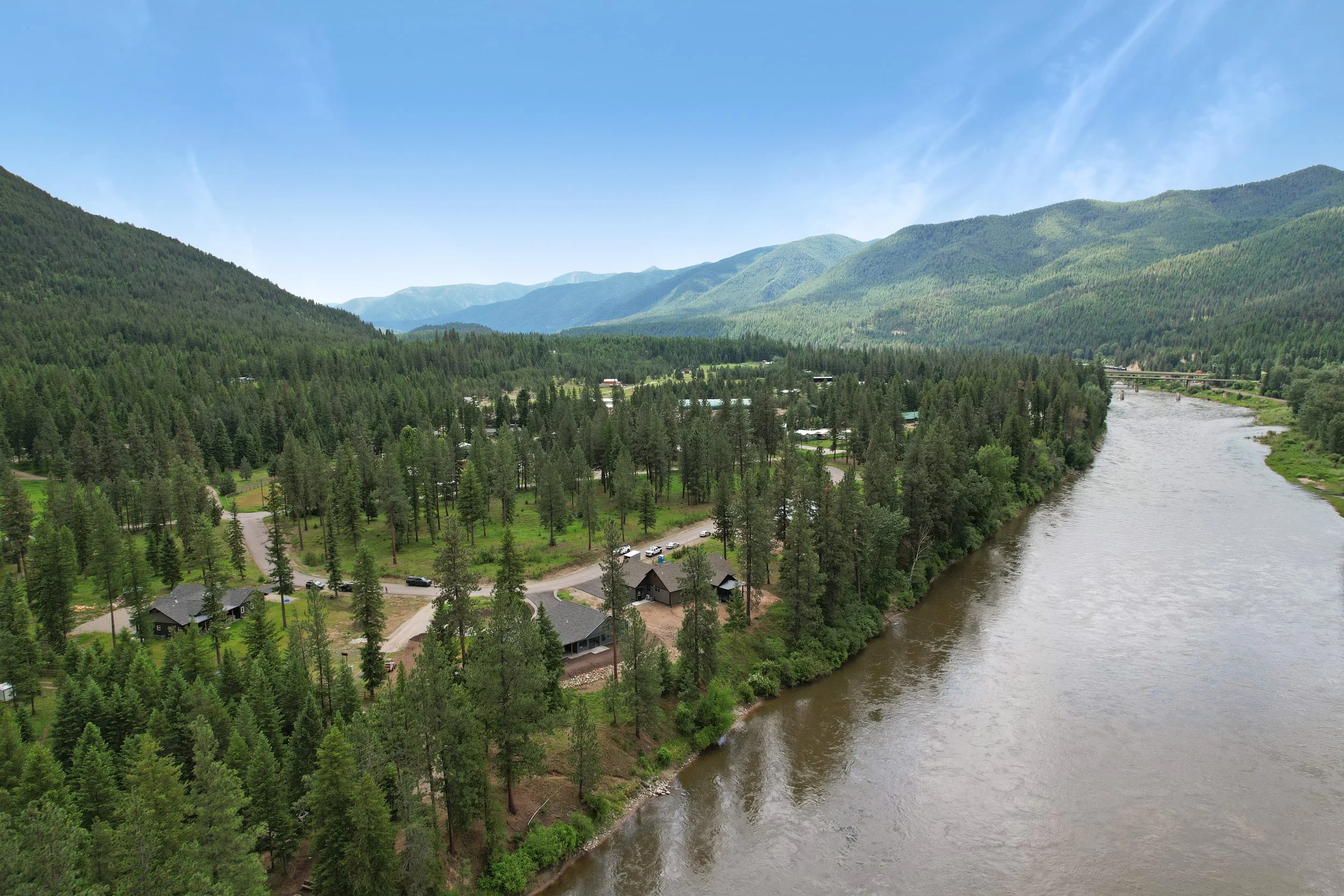 A scenic view of lush green mountains, a river flowing through a forested valley, and a small residential area with houses and cars beneath a clear blue sky.