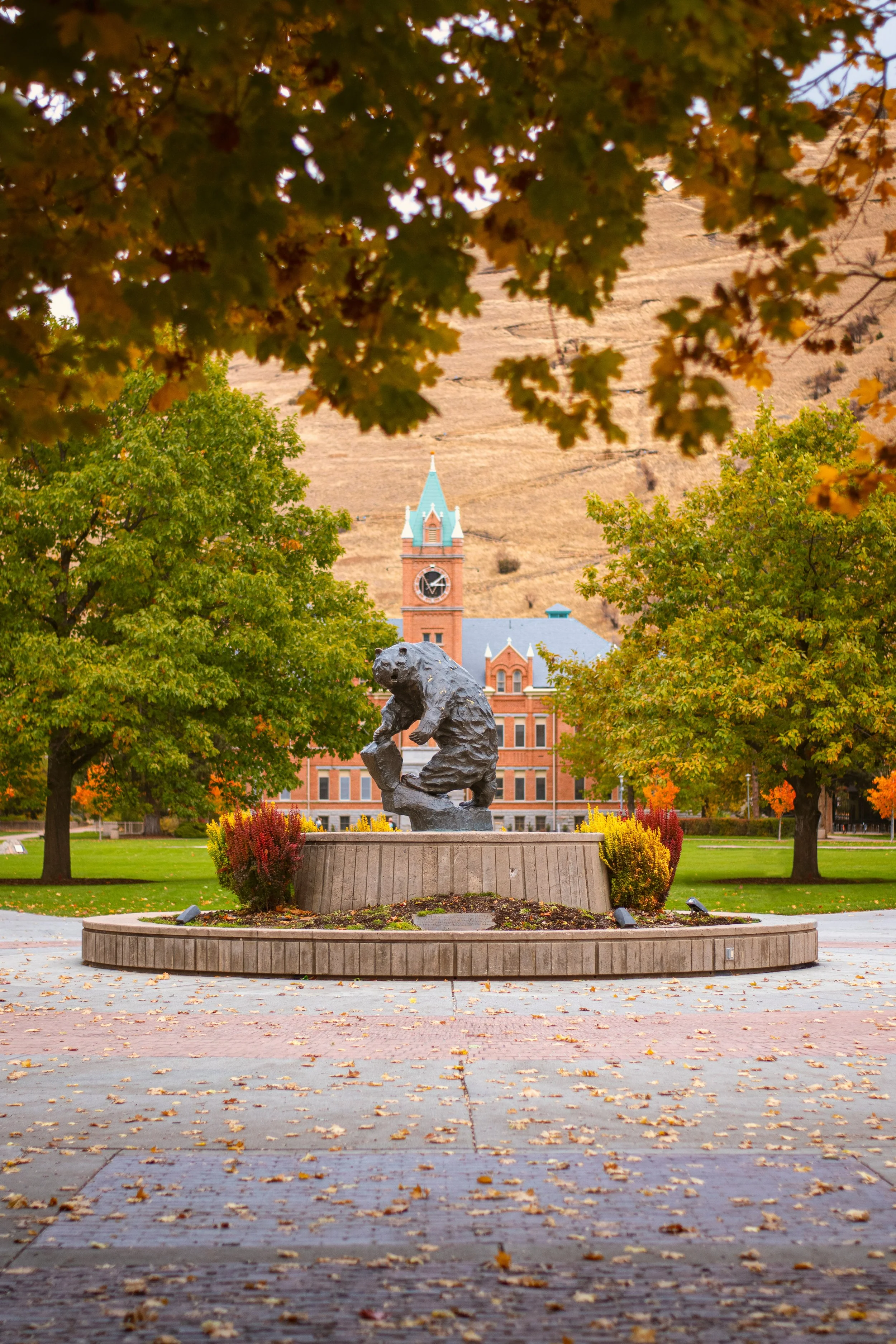 A park scene in autumn with a bronze bear statue in the foreground on a circular stone platform. Surrounding the statue are trees with green, yellow, and orange leaves, with some fallen leaves on the brick-paved walkway. In the background, there's a historical brick building with a clock tower and a turquoise spire, and a light brown hill with sparse vegetation.