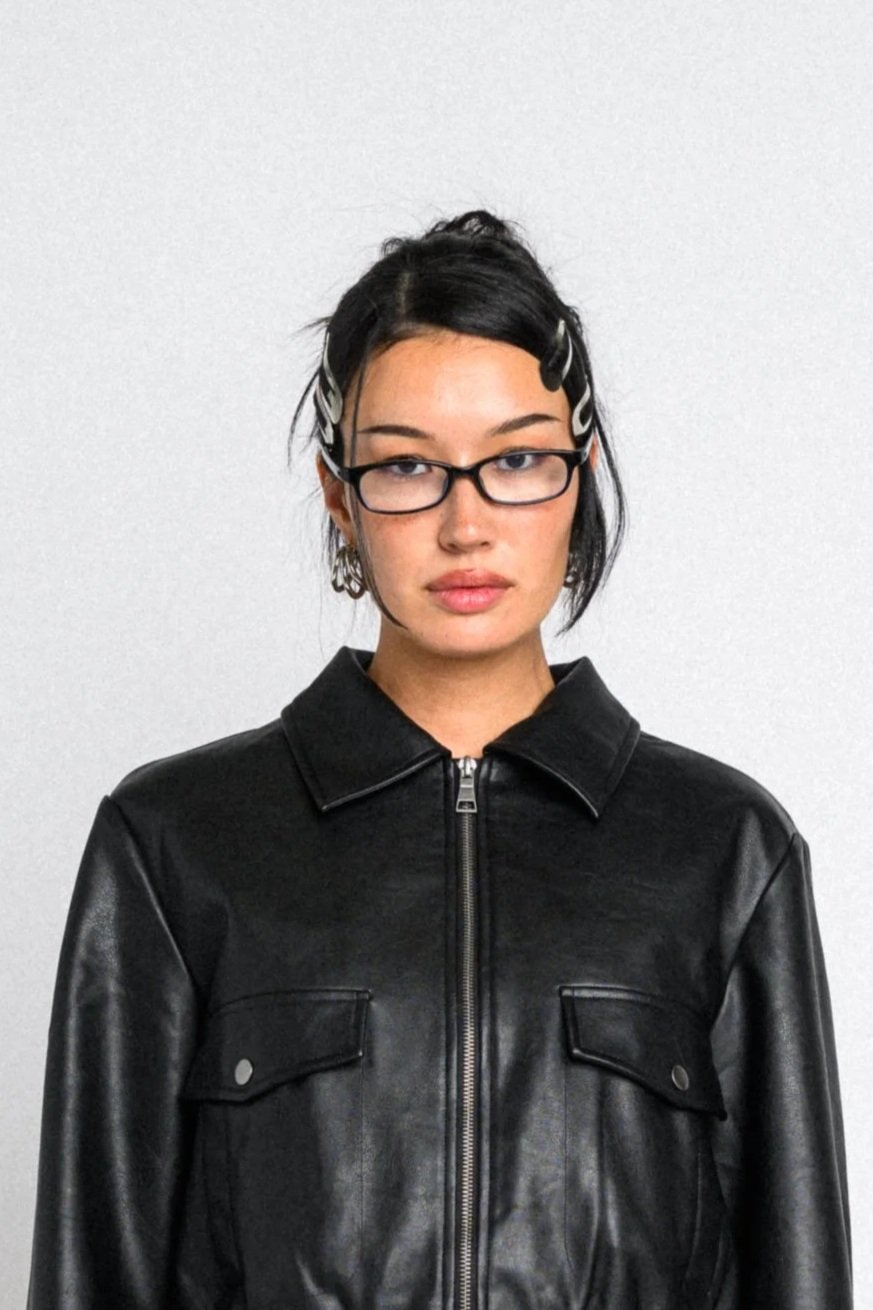 Woman with short black hair, glasses, and hair clips, wearing a black leather jacket, standing against a plain white wall.