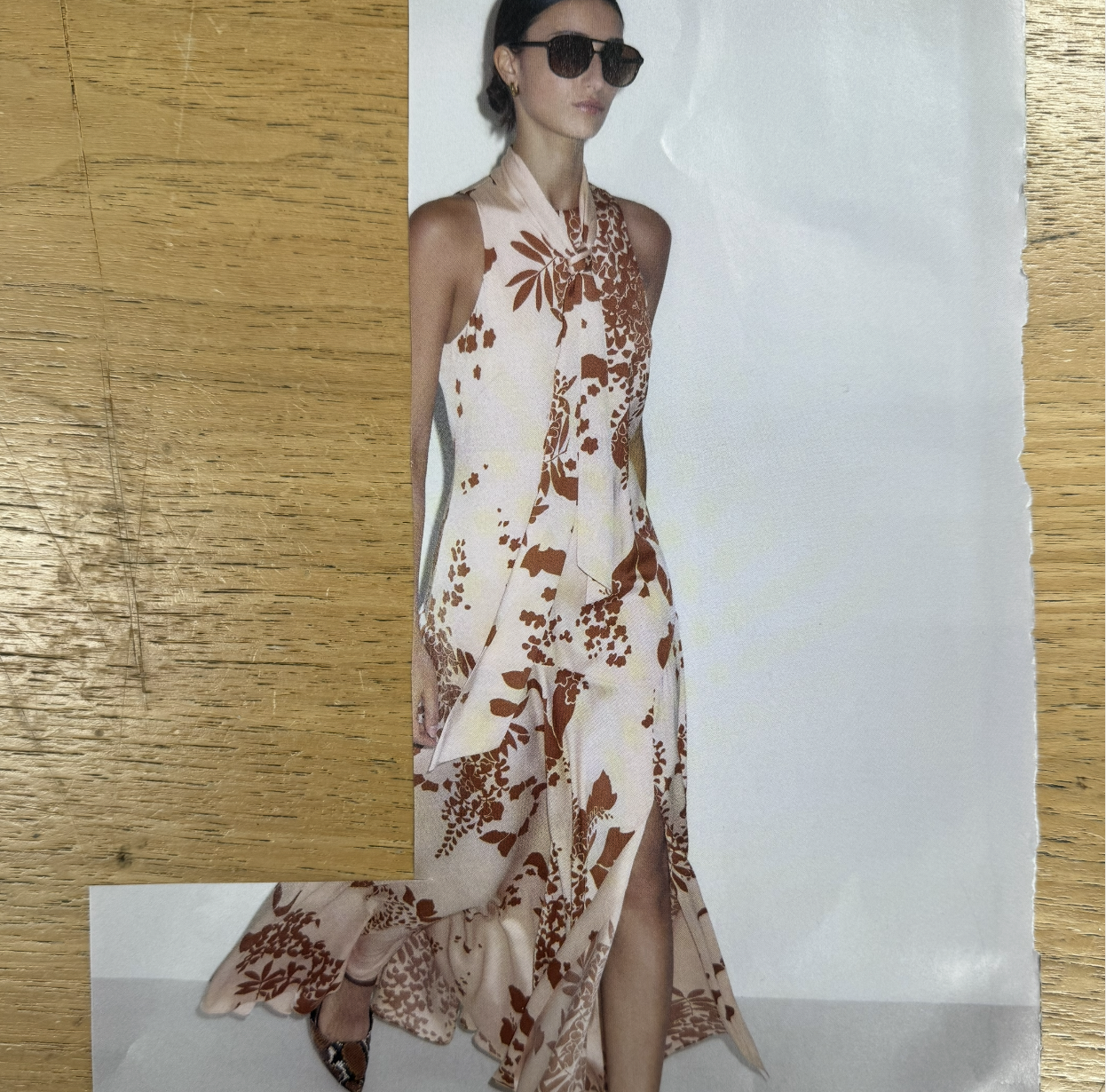 A woman wearing dark sunglasses, a beige and brown patterned sleeveless dress with a slit, and earrings, standing against a plain background.